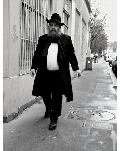 A bearded man in traditional Jewish attire, including a long black coat and hat, walking on a city sidewalk. The street is lined with buildings and trees. The image is in black and white.