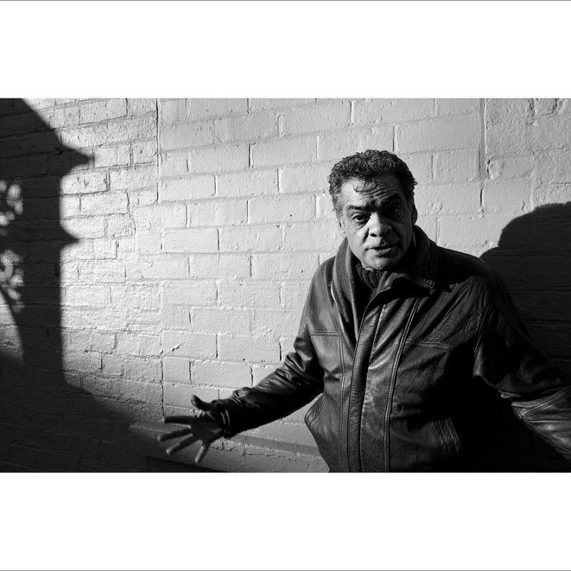Black and white photo of a man in a leather jacket standing against a brick wall with shadows.