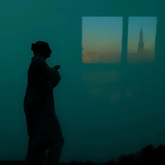Silhouette of a person with arms crossed against a blue-green background; a window with a view of a church spire at sunset visible.