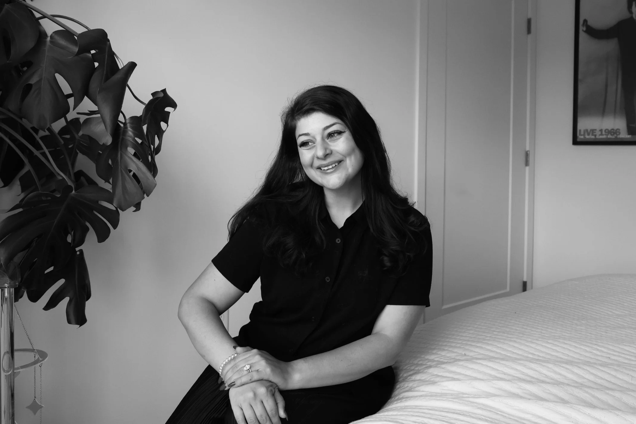 A smiling woman with dark hair sitting on a hospital or clinic bed, wearing a dark shirt, with a large leafy plant nearby, and a poster on the wall in the background.