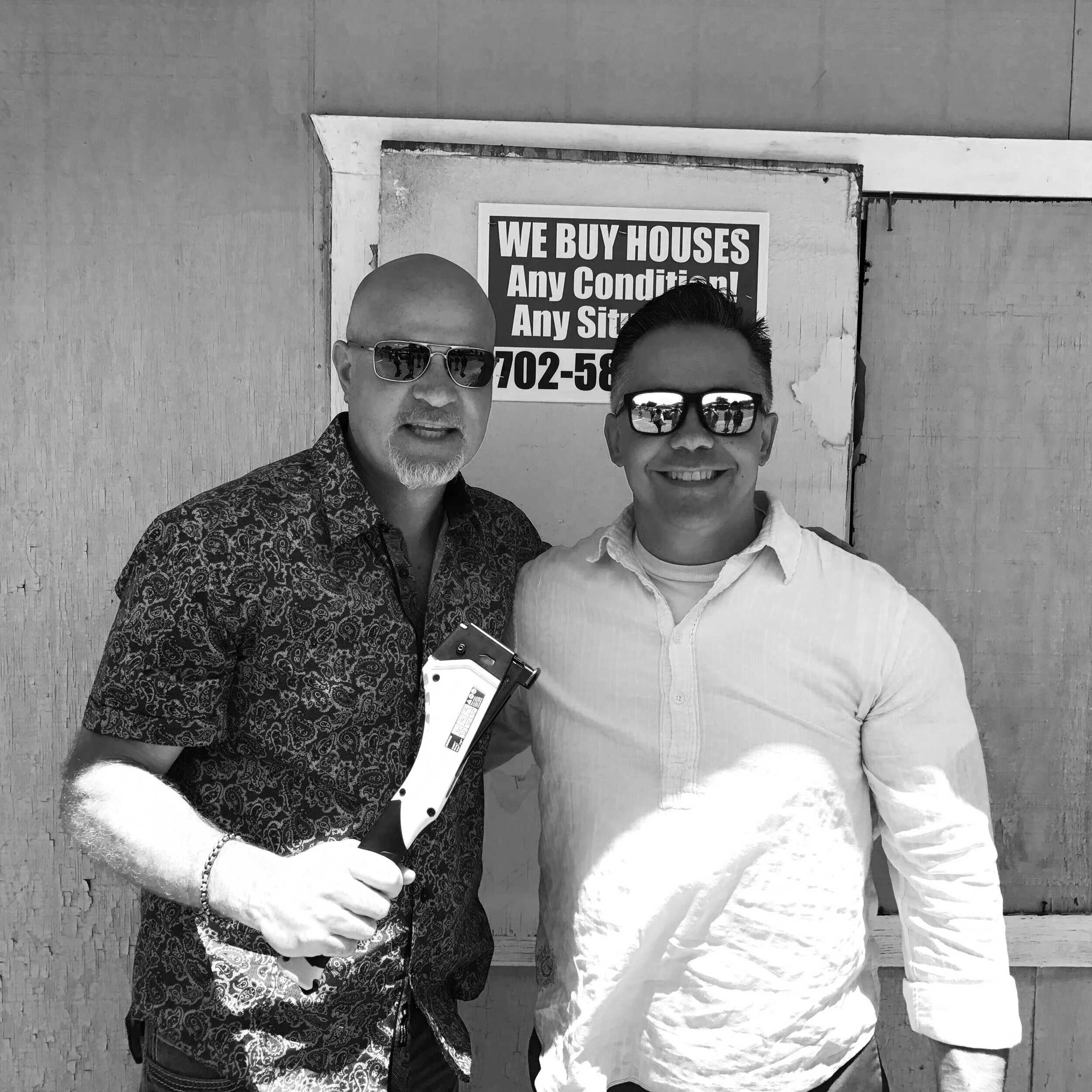 Two men wearing sunglasses, smiling and standing side by side outdoors in front of a sign that reads "We Buy Houses" and a wooden wall. One man is holding a pair of bolt cutters.