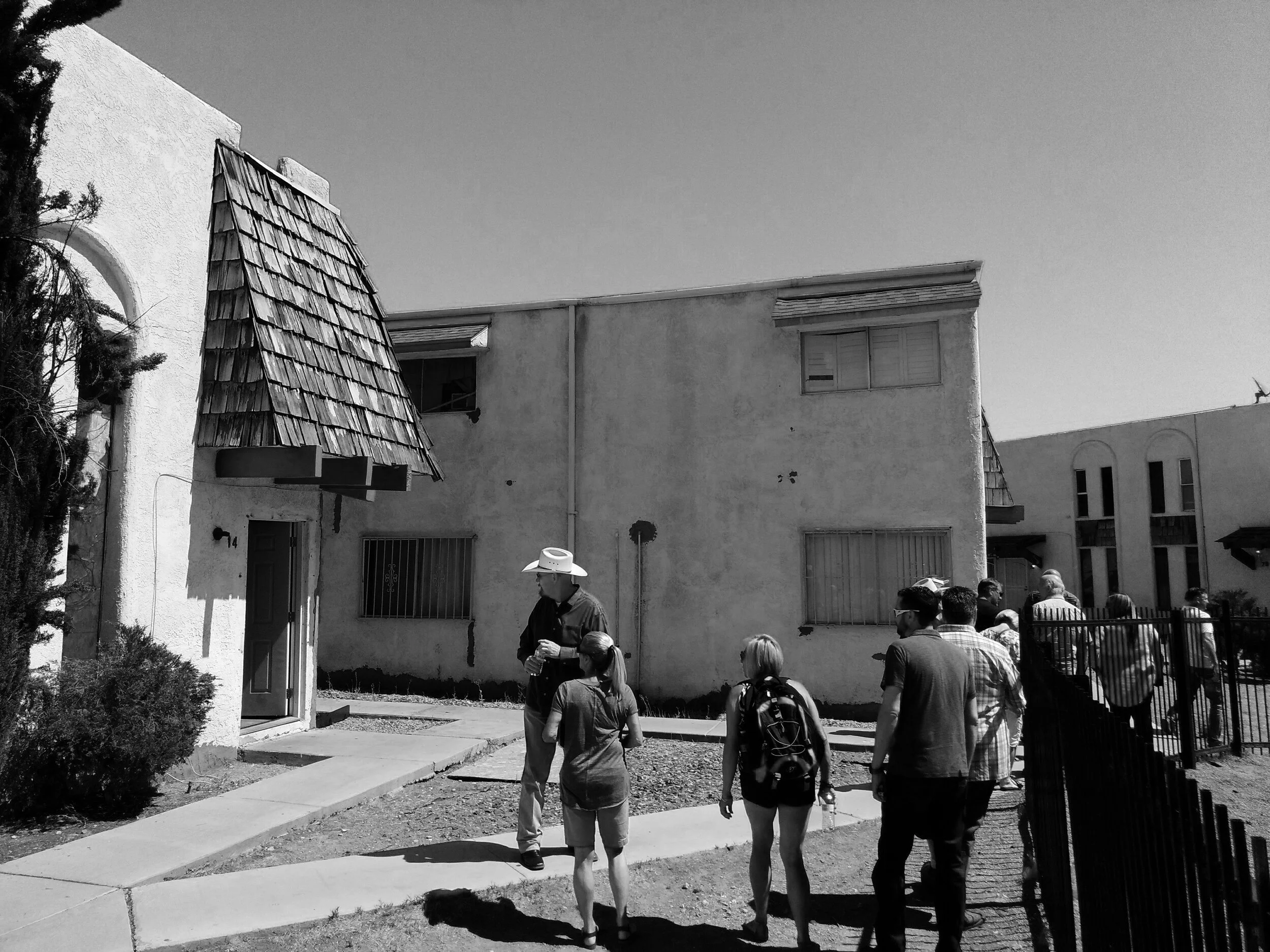 A group of people, including a man with a cowboy hat, walking past a building with stucco walls and small, shield-shaped awnings over the windows, under a clear sky.