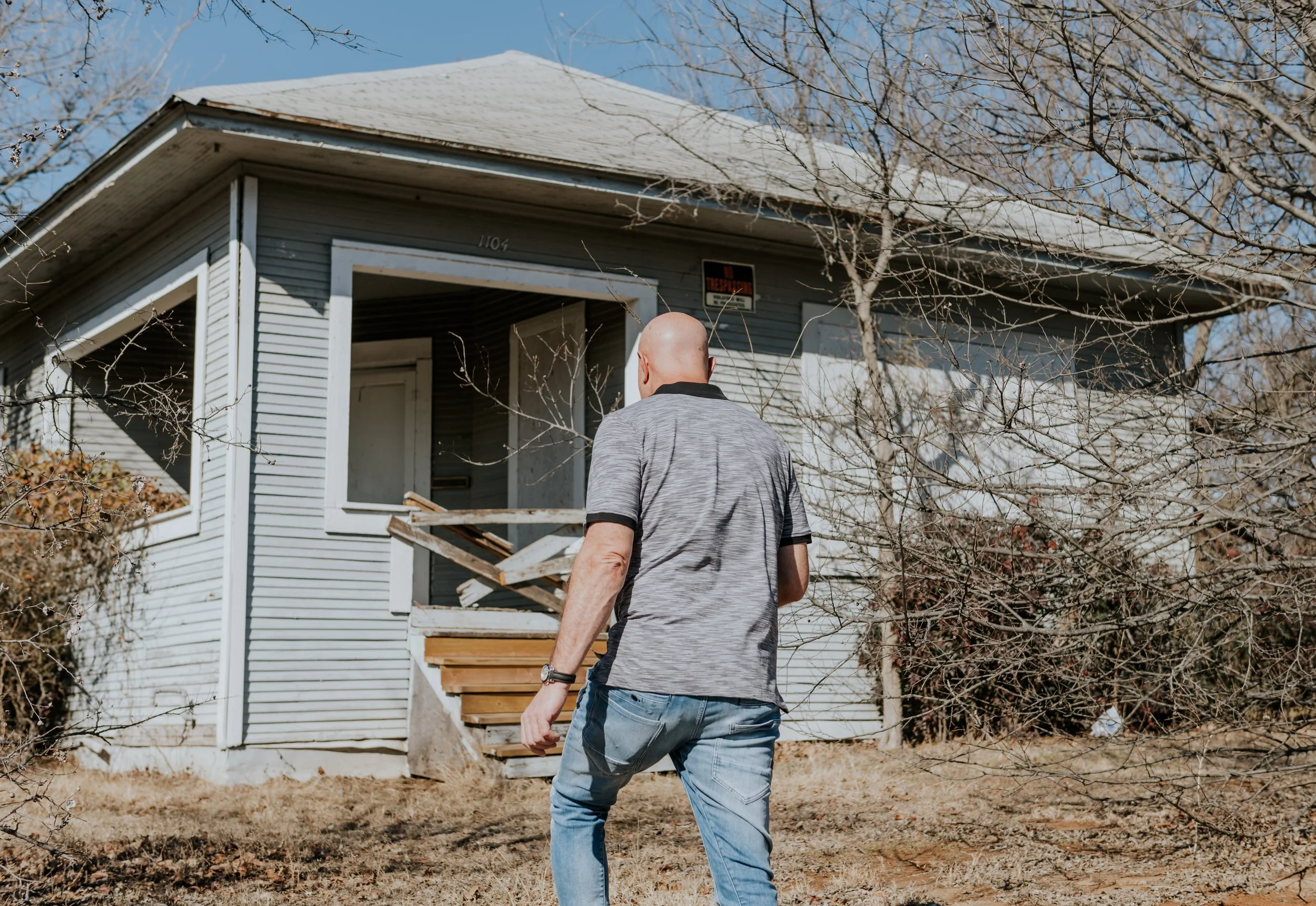 A man with a shaved head wearing a gray polo shirt and jeans walking toward an abandoned house with a broken front porch, surrounded by leafless trees and dry grass.