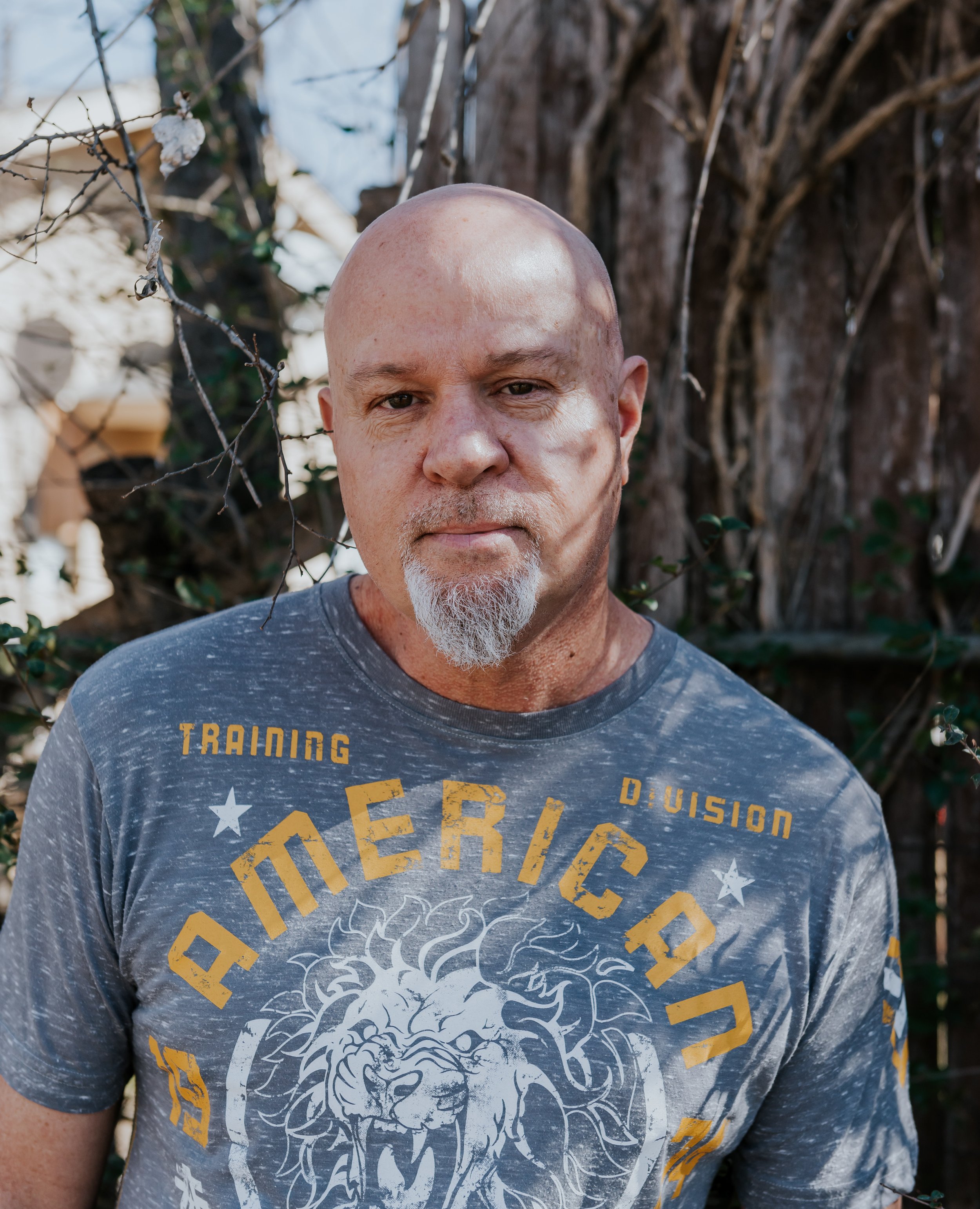 A man with a goatee and shaved head stands outdoors in front of a tree and dry vines, wearing a gray Arizona Dvision t-shirt with a lion graphic and yellow text.