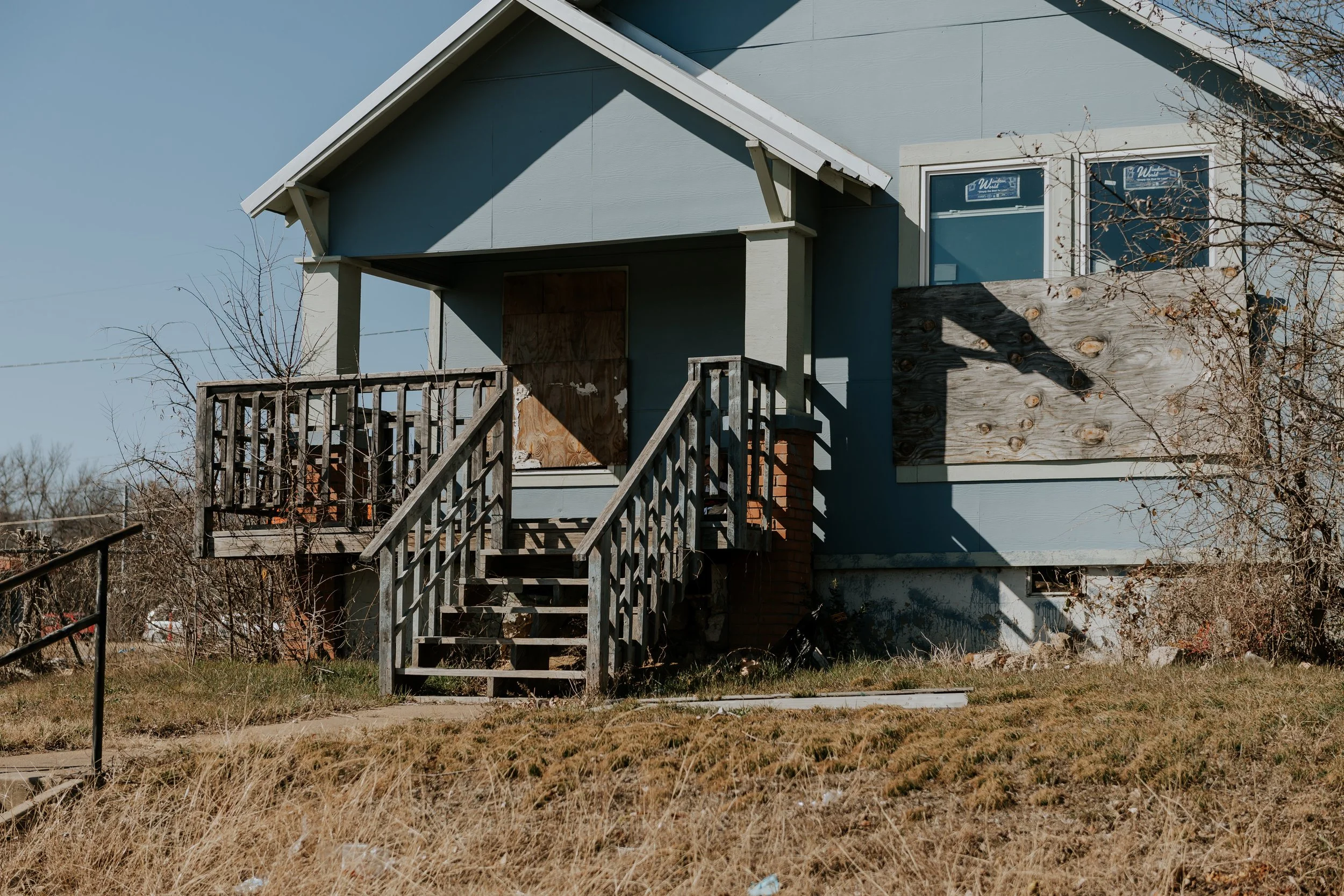 An abandoned house with a wooden staircase leading to a boarded-up front door and a boarded-up window on the second floor. The house has light blue siding, and the yard is overgrown with dry grass and some bushes with no leaves.