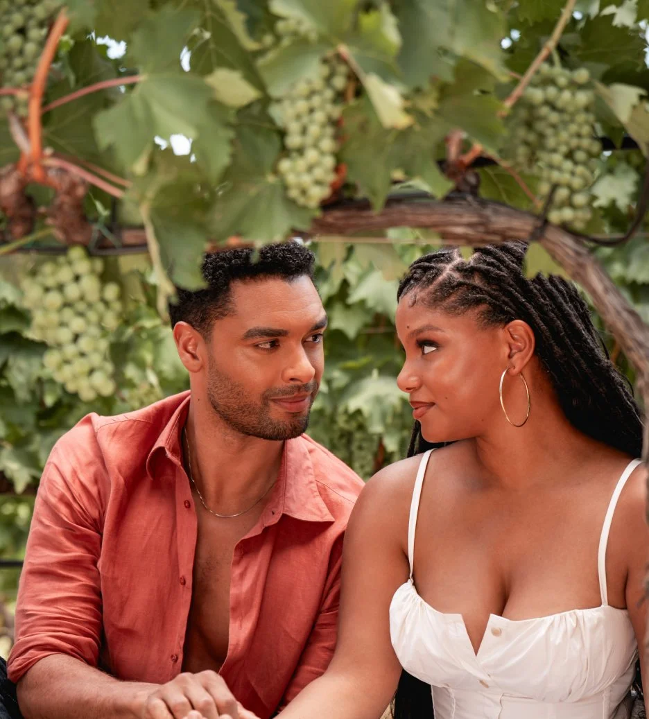 Halle Bailey and Rege Jean Page sit under grape vines looking into each others eyes.