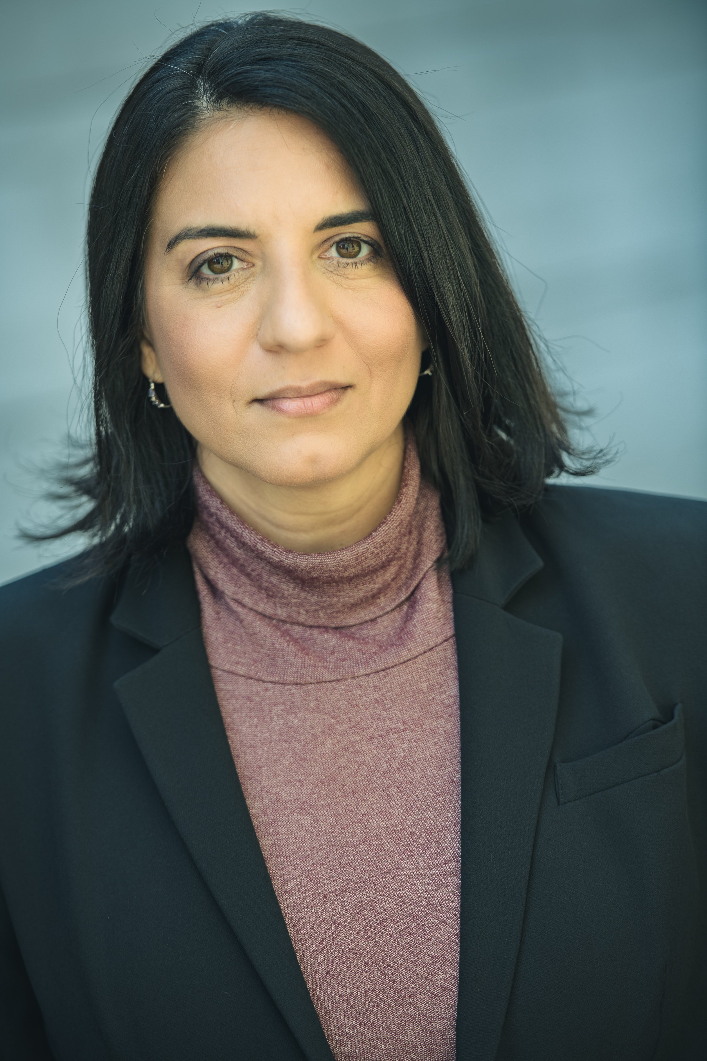A woman with shoulder-length black hair, wearing a dark blazer and a maroon turtleneck sweater, looking at the camera with a neutral expression. The background is a plain gray color.