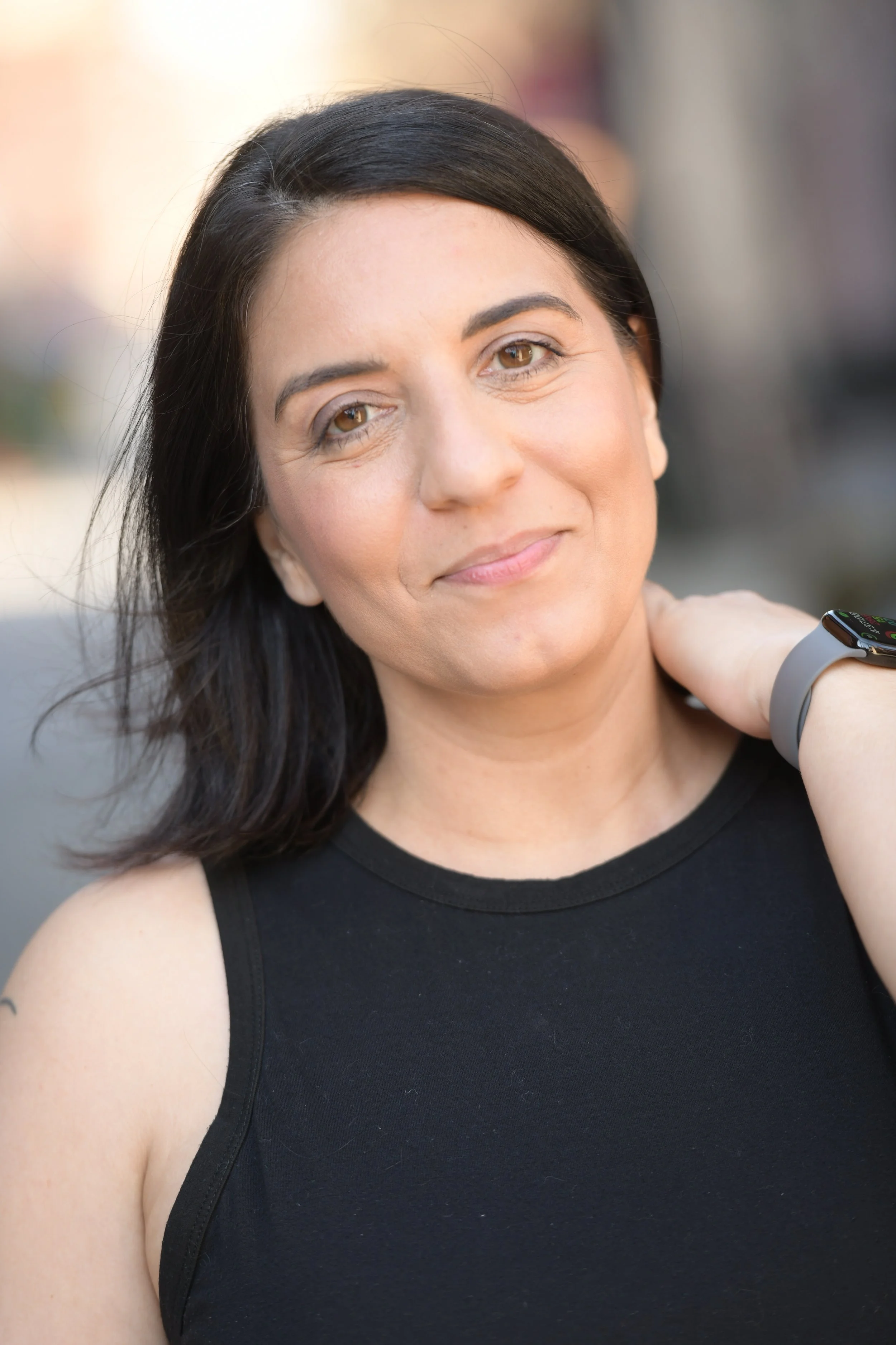 Close-up of a woman with dark hair and light skin wearing a black sleeveless top, smiling softly, with a smartwatch on her wrist, outdoors with blurred background.