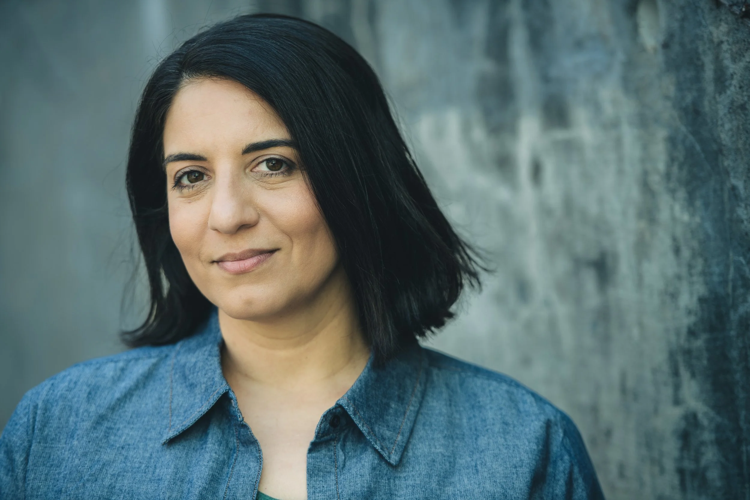 A young woman with black hair, wearing a blue denim shirt, standing against a gray textured wall.