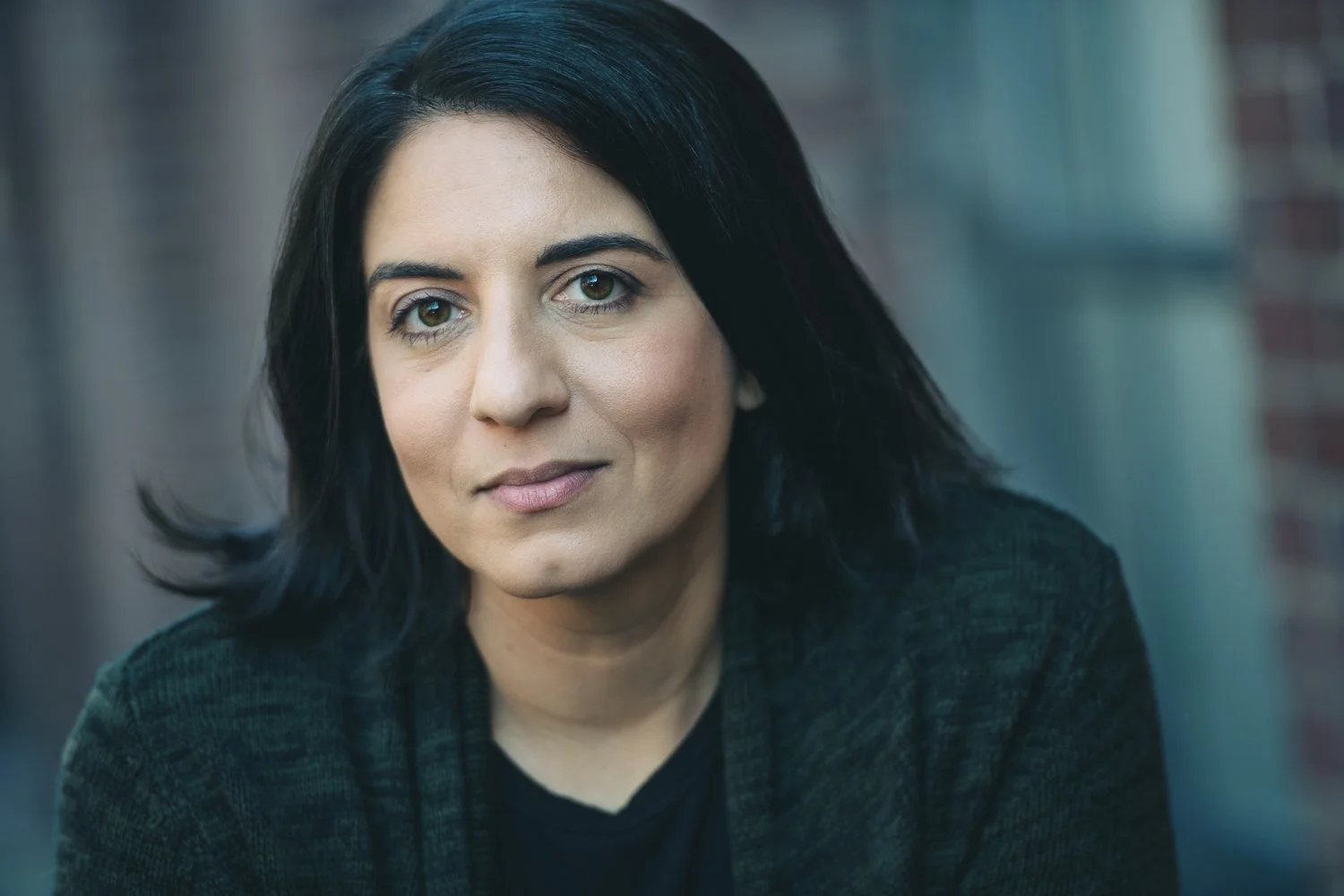 A woman with black hair and light skin looks directly at the camera, wearing a dark jacket, with a blurred background of a brick wall.