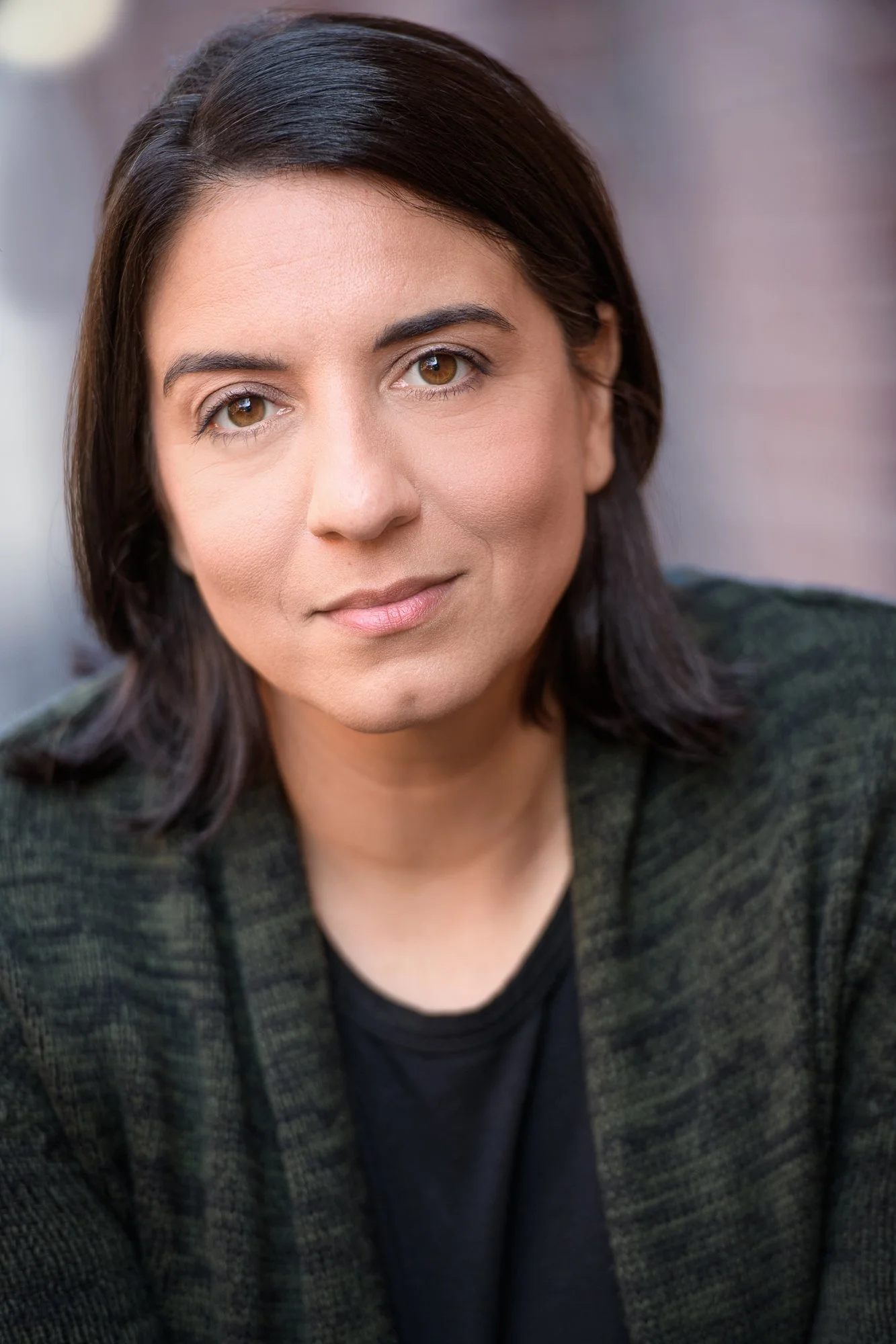 Close-up of a woman with dark brown hair, wearing a dark patterned blazer and black top, looking directly at the camera with a neutral expression.
