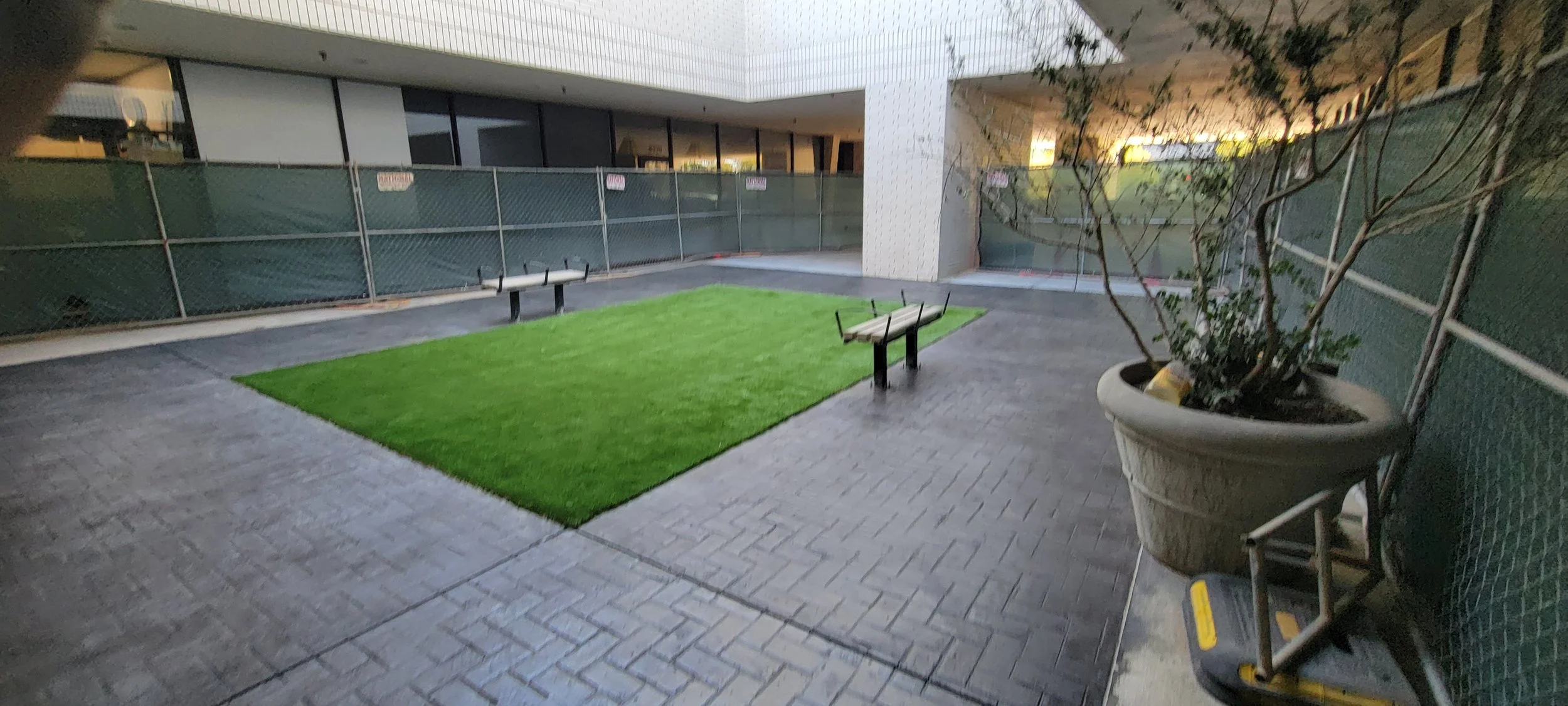 Enclosed courtyard with a rectangular artificial turf area surrounded by patterned paving stones, benches, and a potted tree.