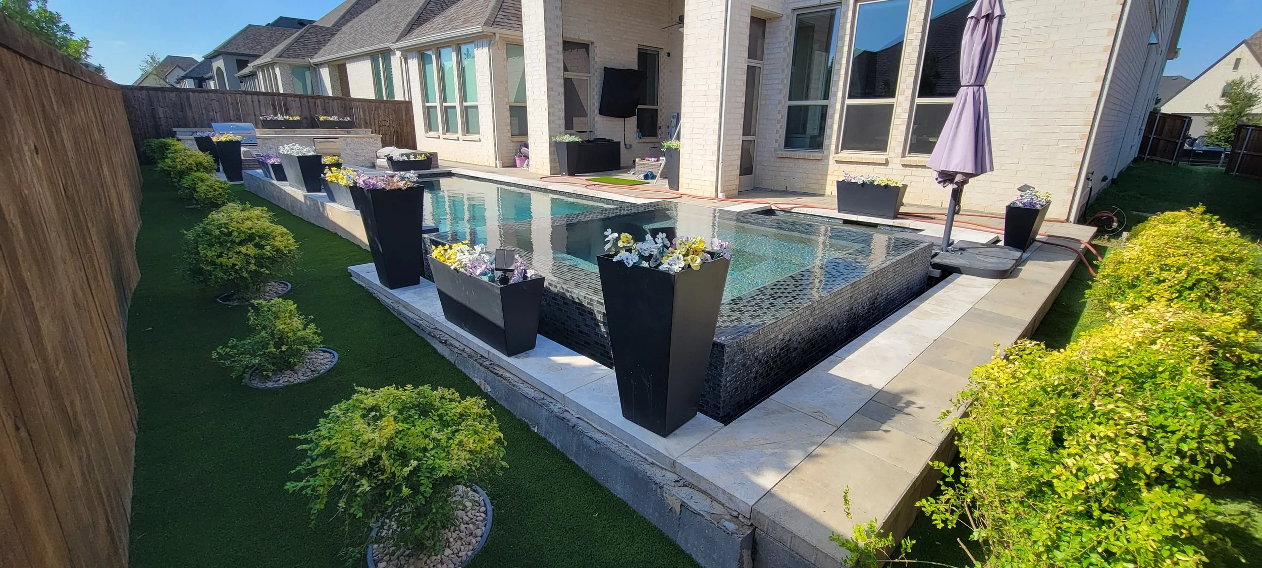 Professionally installed artificial turf surrounds a backyard swimming pool and patio beside a brick house.