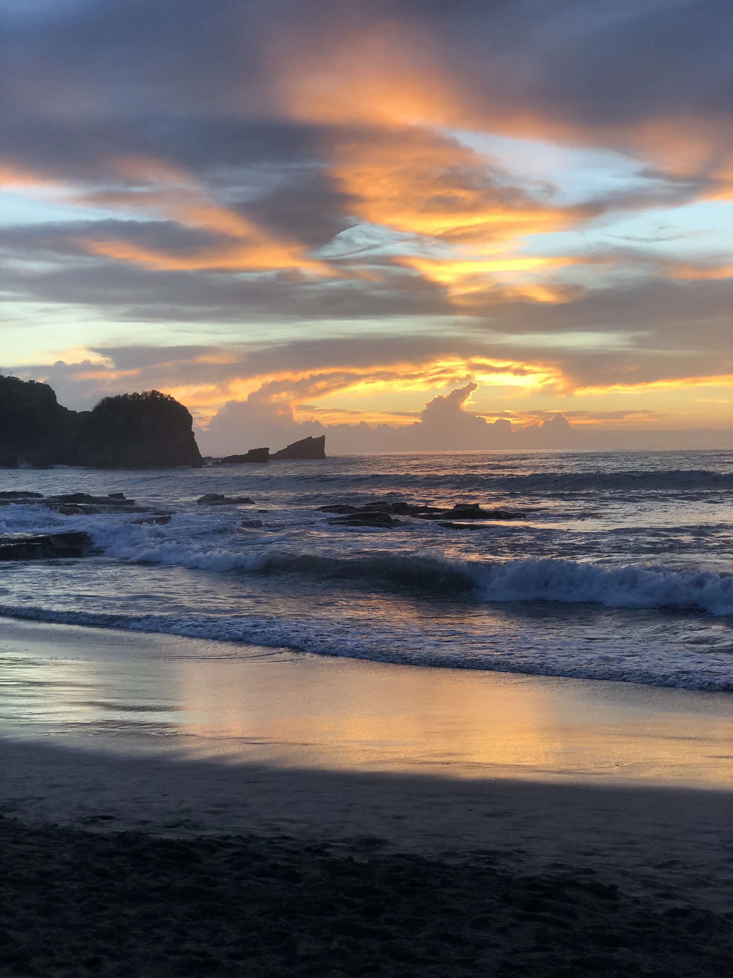 Sunset over the ocean with a partly cloudy sky, reflecting orange and blue hues on the water and shoreline, with rocky formations in the distance.