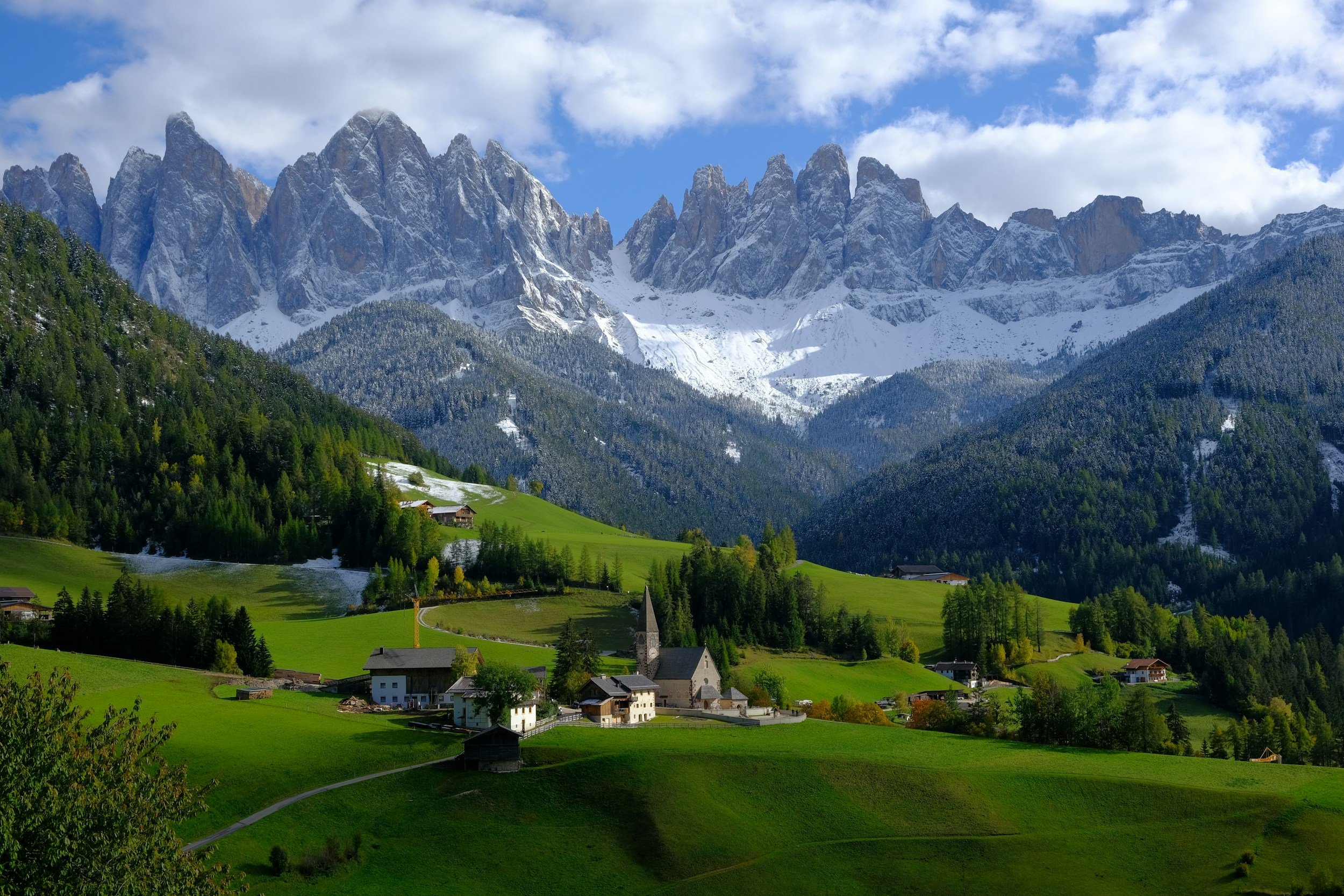 Scenic view of a small village with a church, houses, and lush green fields, situated in the foothills of snow-capped mountains under a partly cloudy sky.