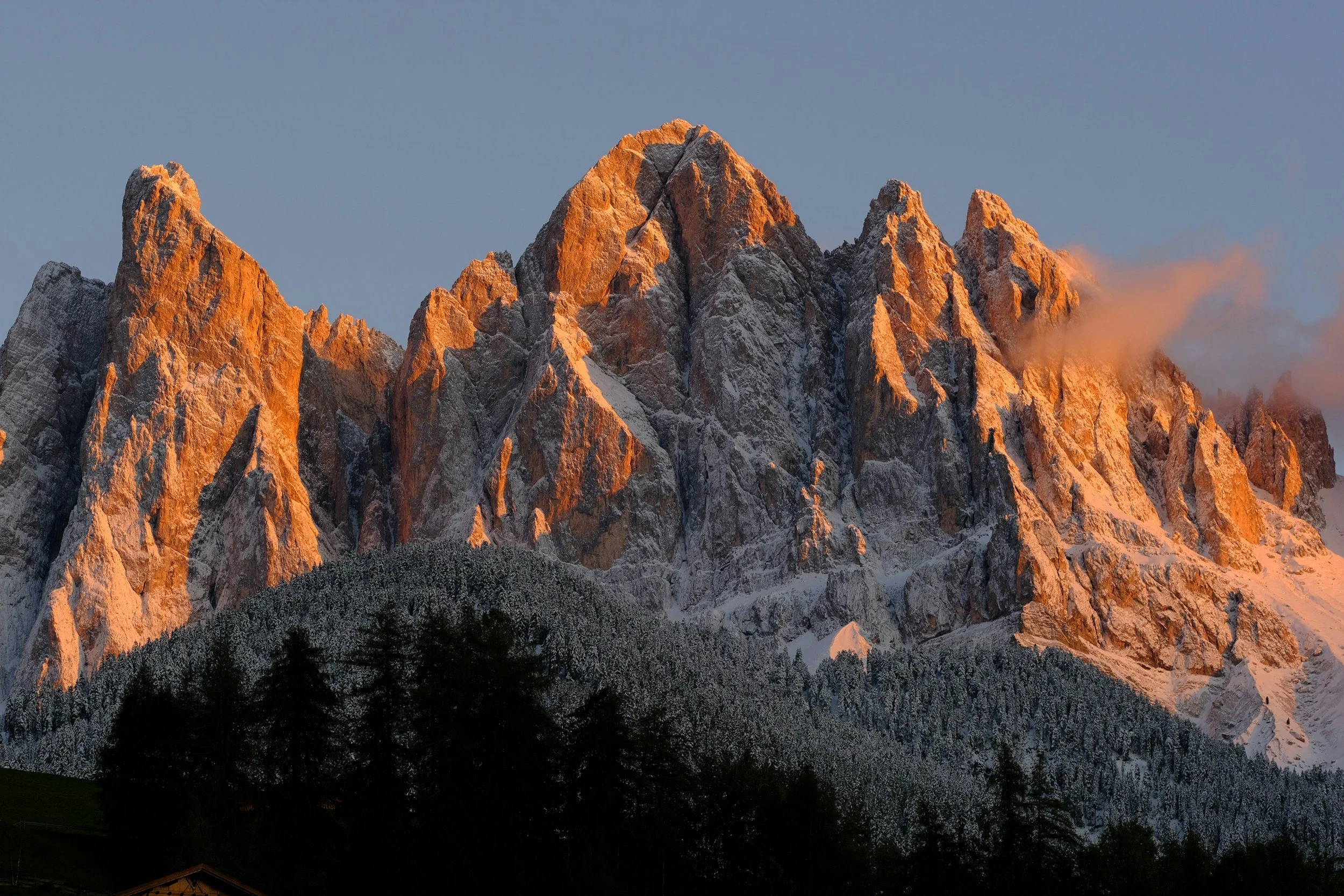 Sunlit mountain peaks during sunset with snow and forested lower slopes.