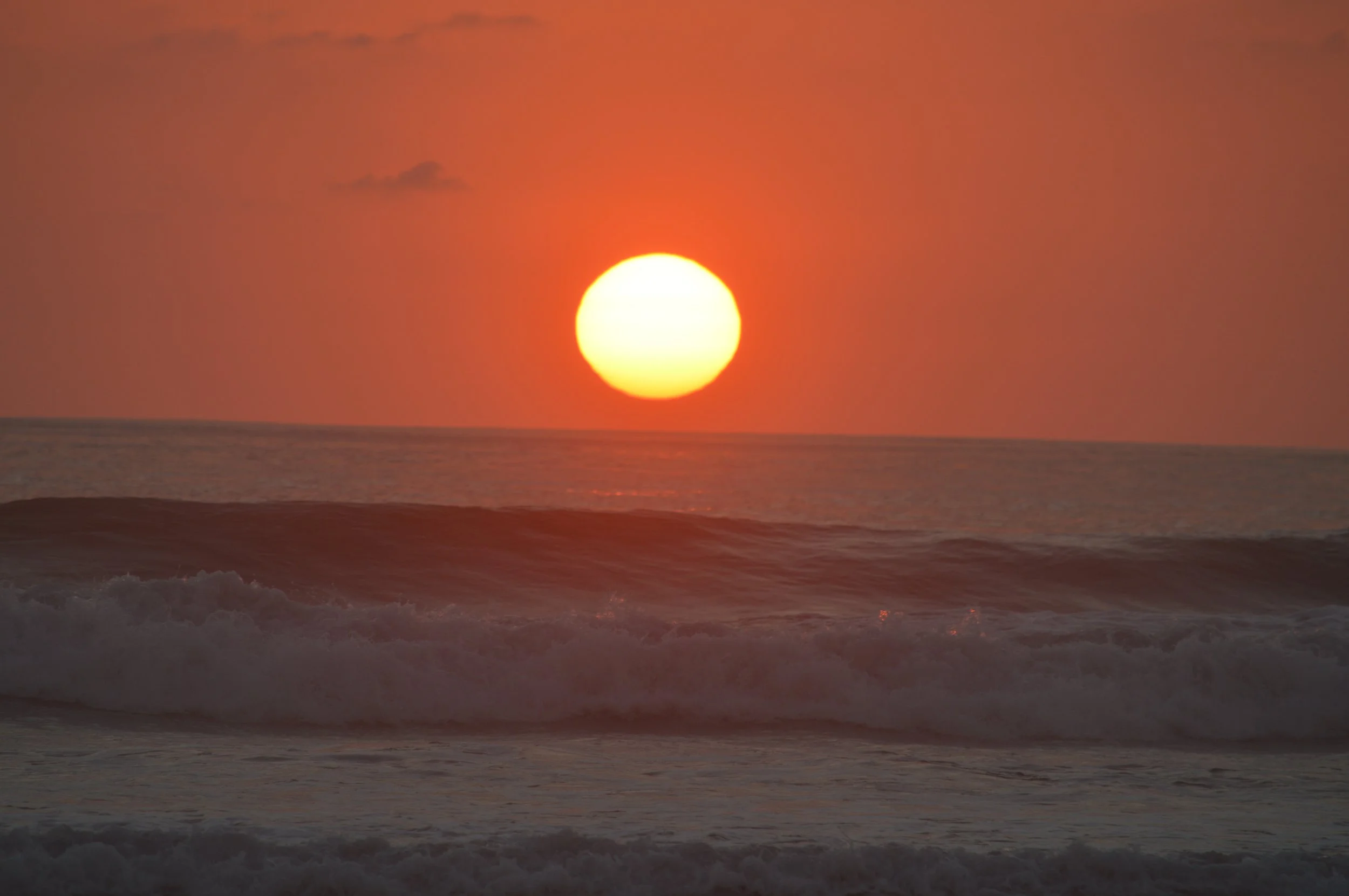 Sunset over the ocean with waves in the foreground and a mostly clear sky with a few clouds
