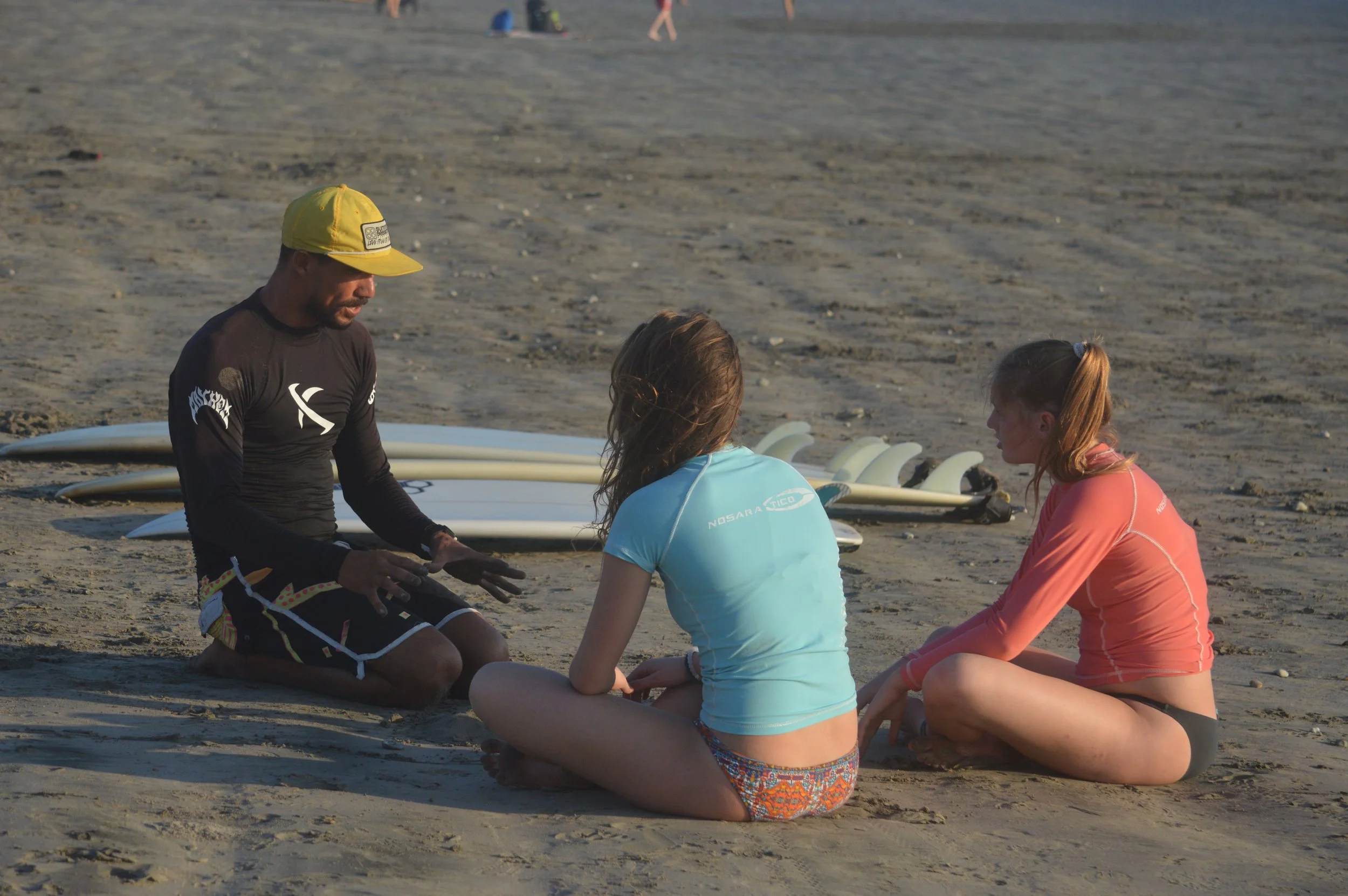 A man kneeling on the beach talking to two women sitting cross-legged. Surfboards are lying on the sand behind them.