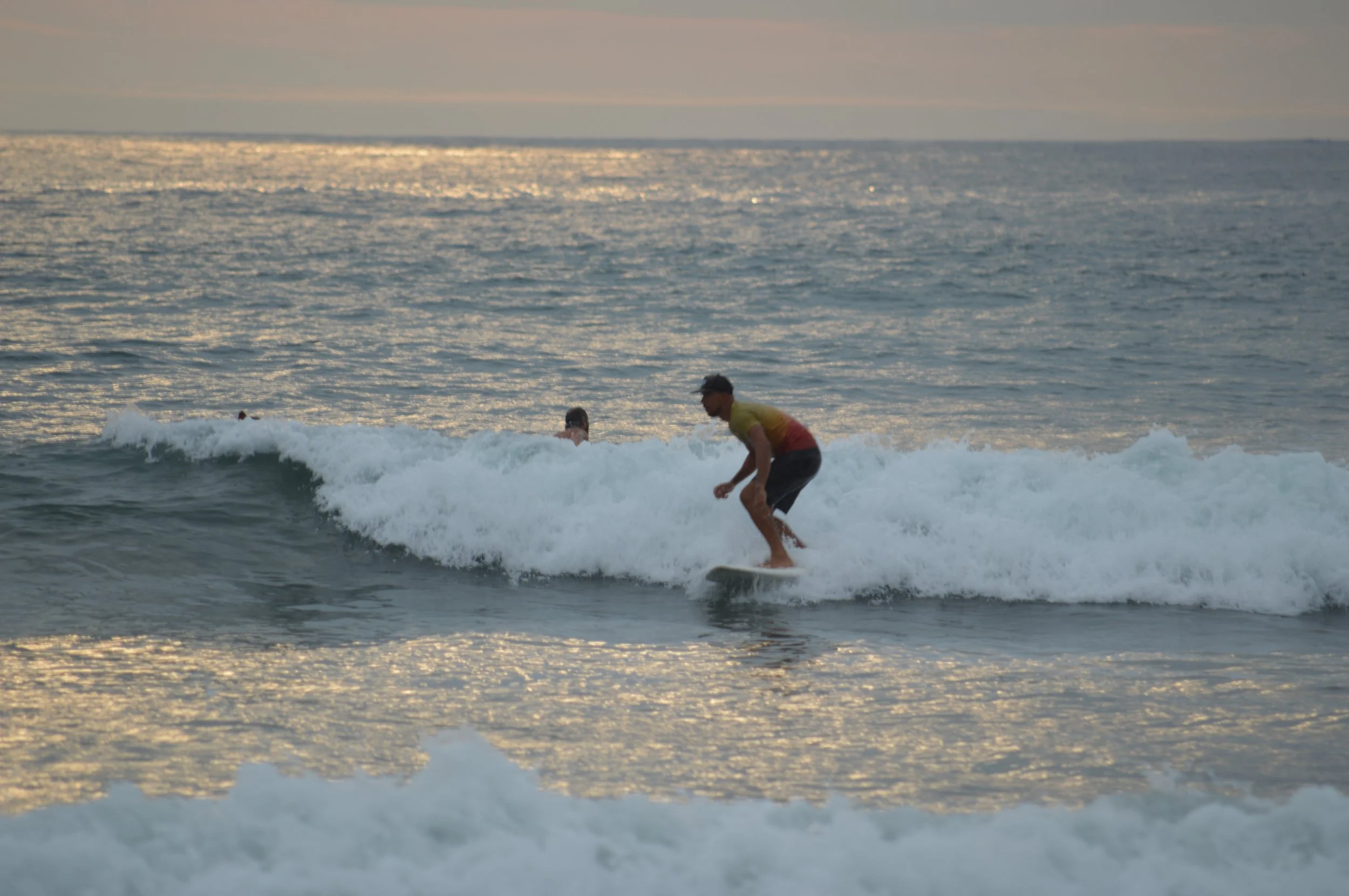 A person surfing on a small wave in the ocean during sunset or sunrise, with two other swimmers in the background and the shimmering water reflecting the sky.