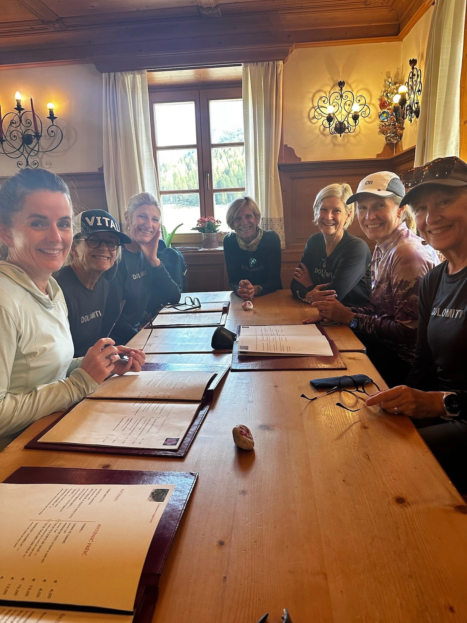 Group of women sitting at a wooden table in a cozy, wood-paneled room with large windows and decorative wall sconces.