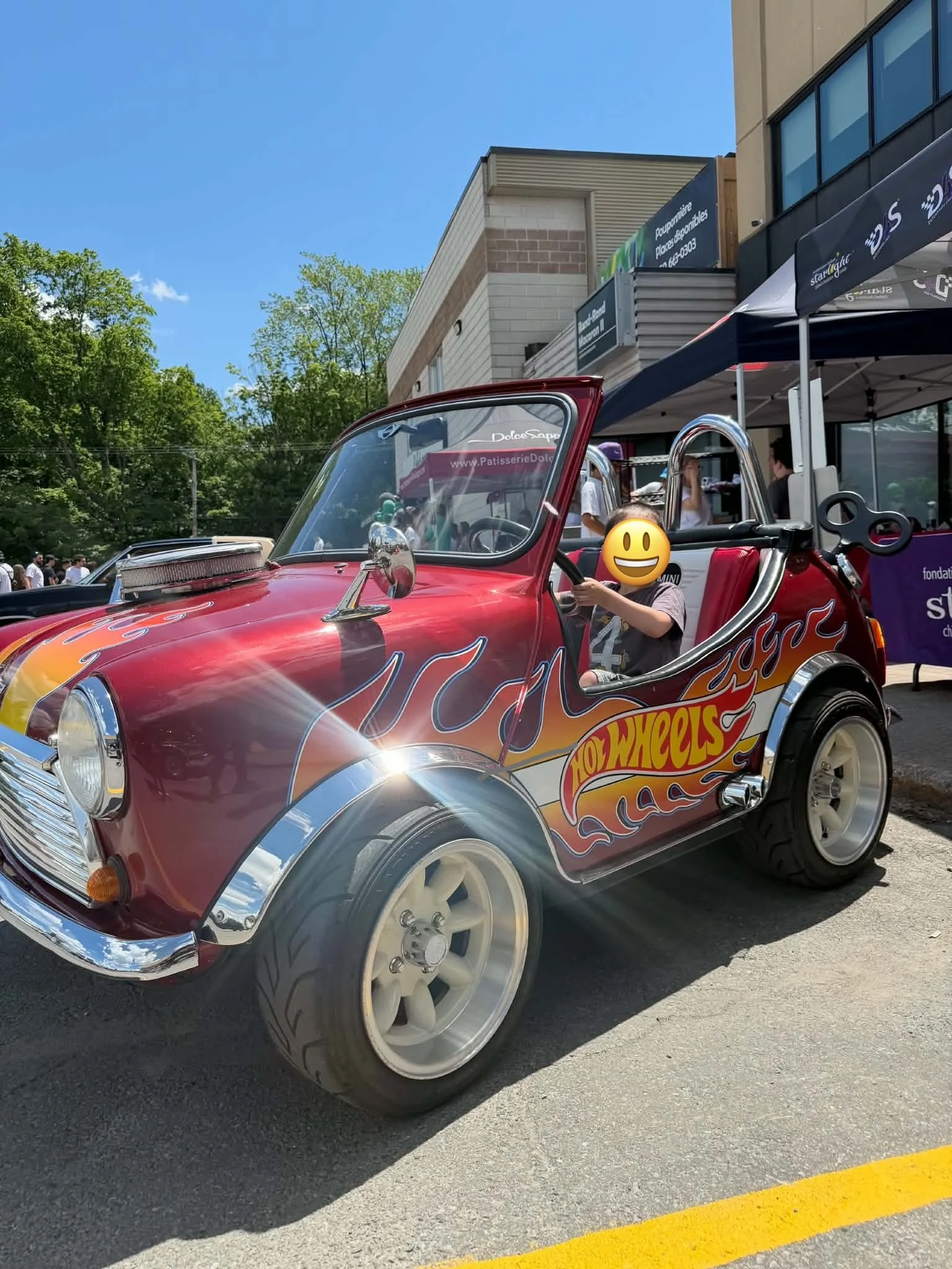 A red Hot Wheels toy car modeled after a vintage vehicle, parked outdoors on a parking lot with trees and buildings in the background, featuring a child sitting in the driver seat with an emoji face overlay.
