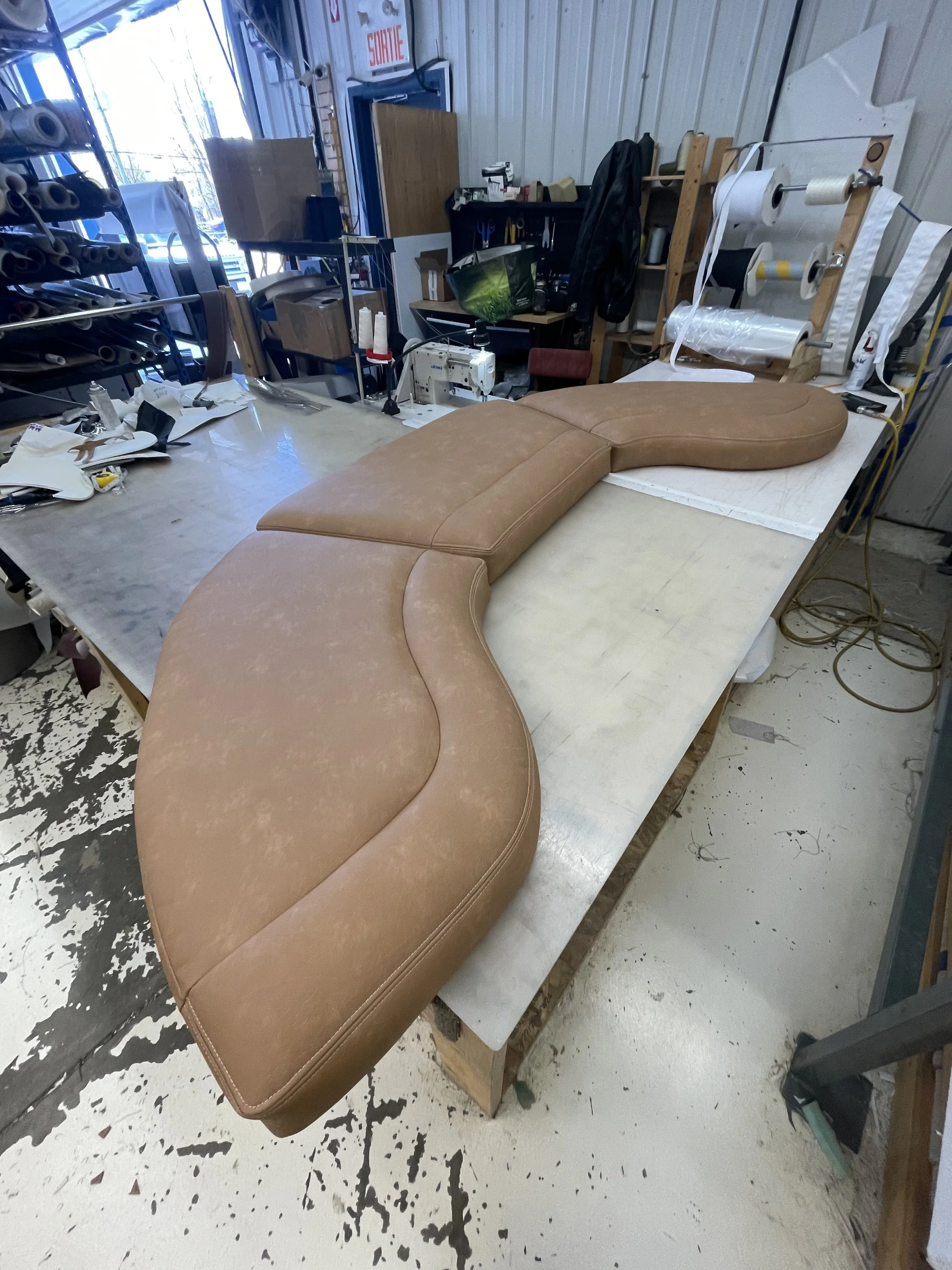 A workspace with a brown cushioned massage or chiropractic table on a white surface, surrounded by various tools, supplies, and equipment in a workshop or repair shop setting.
