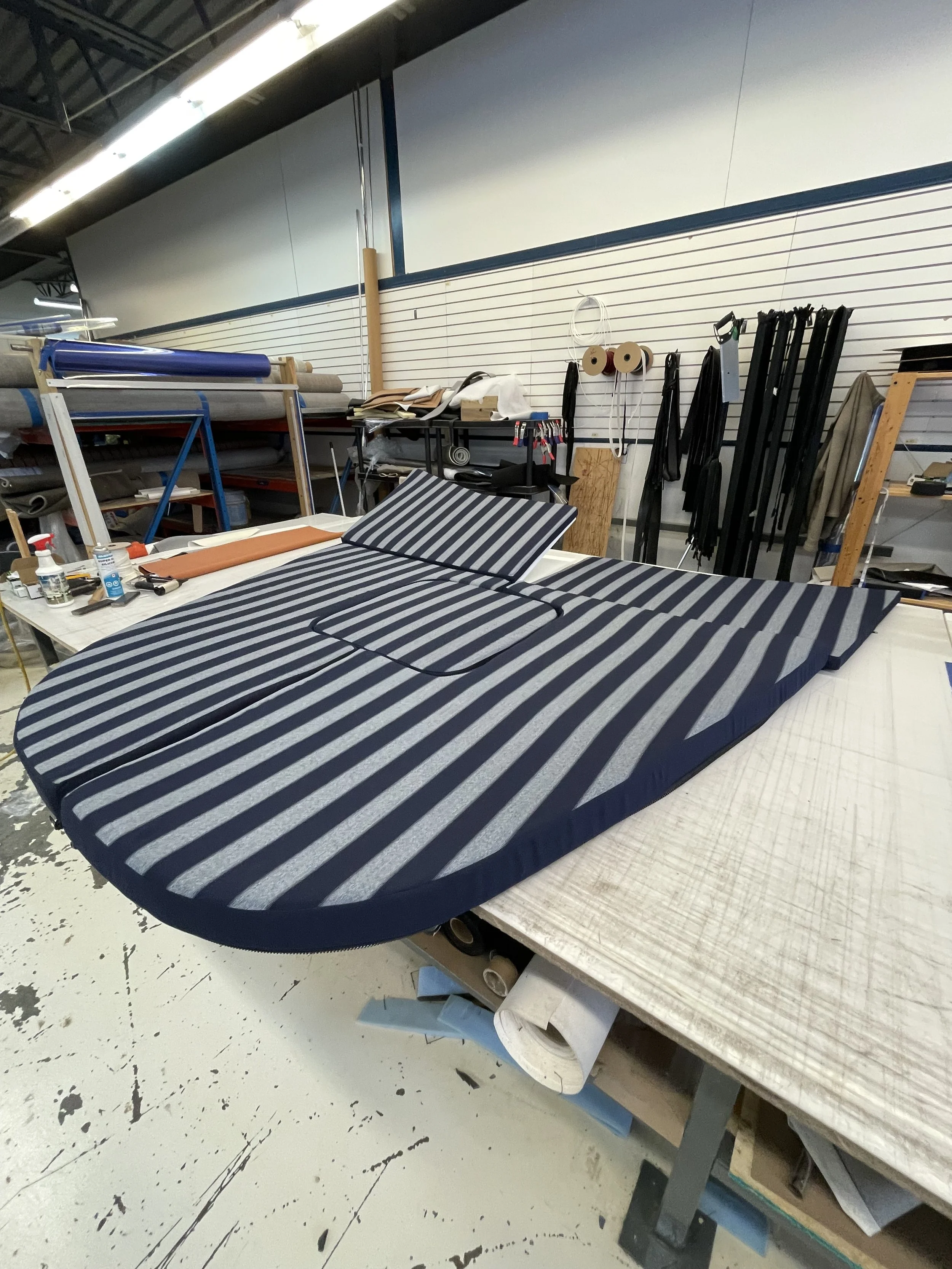 A workspace with a large fabric piece in navy blue and gray stripes spread out on a table, surrounded by tools, materials, and rolls of fabric, with a slat wall in the background holding spools and straps.