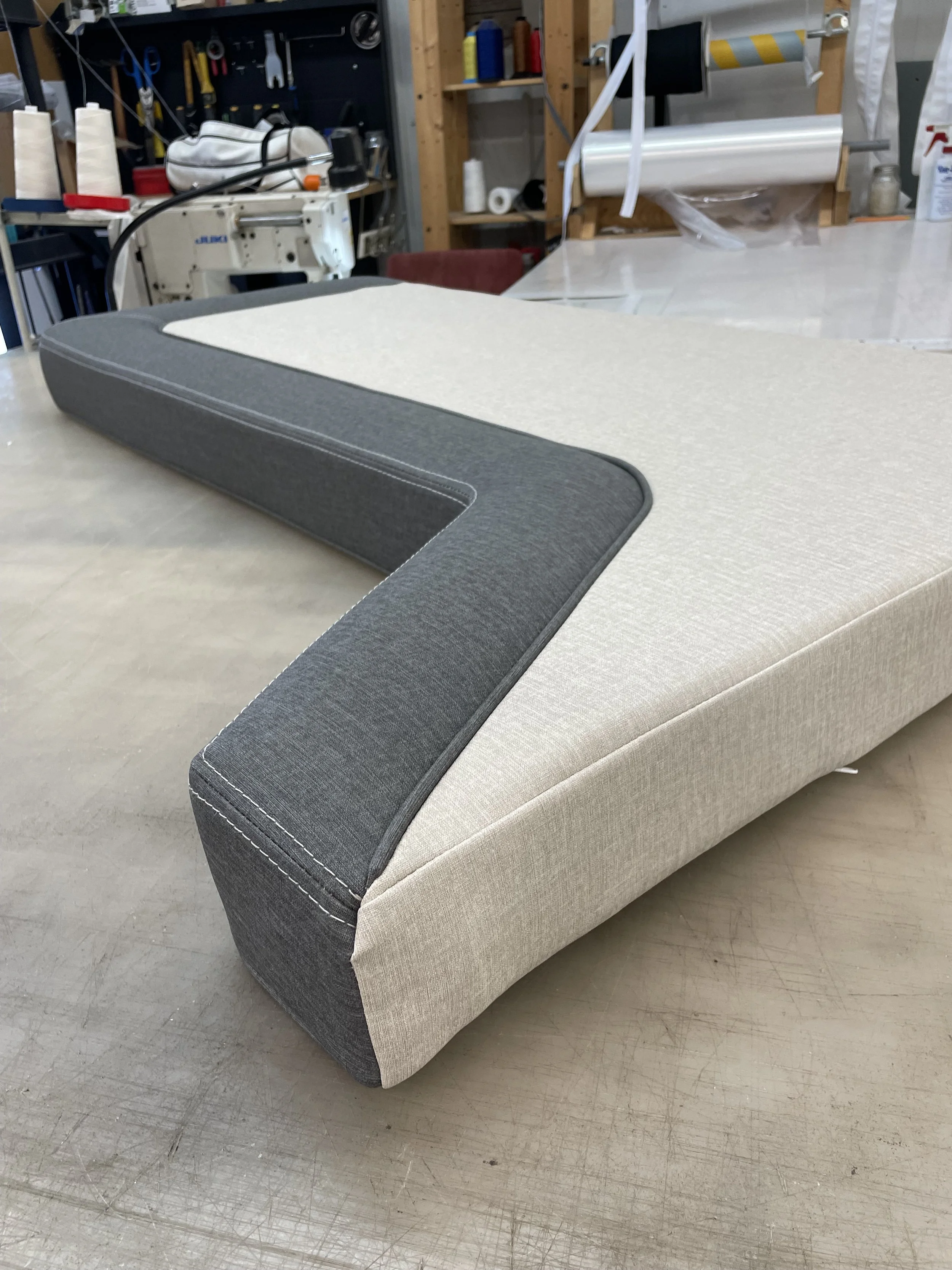A piece of furniture with a beige fabric covering and a gray fabric border on the top edge, placed on a gray concrete floor in a workshop or craft room.