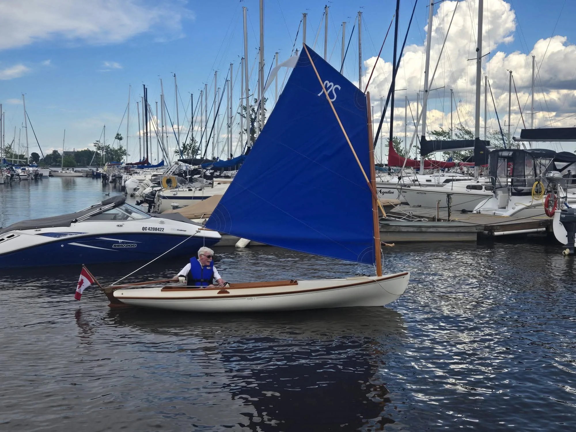 A small wooden sailboat with a person in a life jacket sailing in a marina. The boat has a Canadian flag at the stern and a large blue sail. The marina is filled with numerous larger sailboats and yachts, all docked with tall masts. The sky is partly cloudy with patches of blue visible.