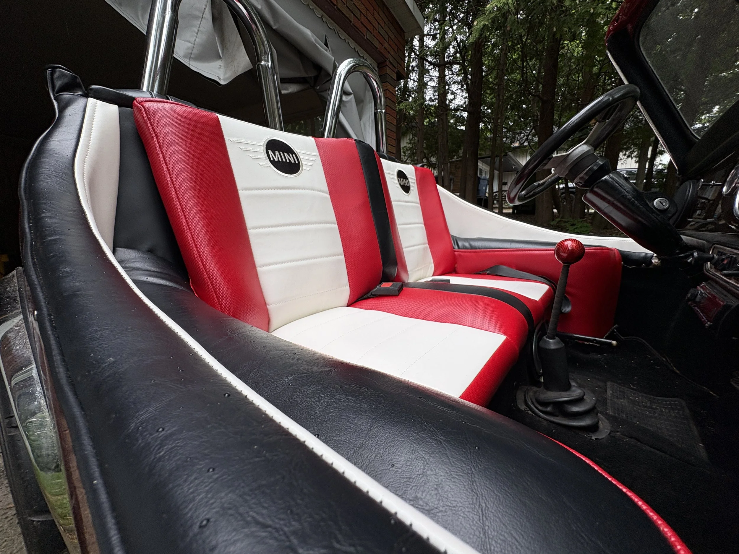 Inside a vintage Mini Cooper car showing the red, white, and black padded seat, black steering wheel, and gear shift lever, with trees and a house visible through the windows.