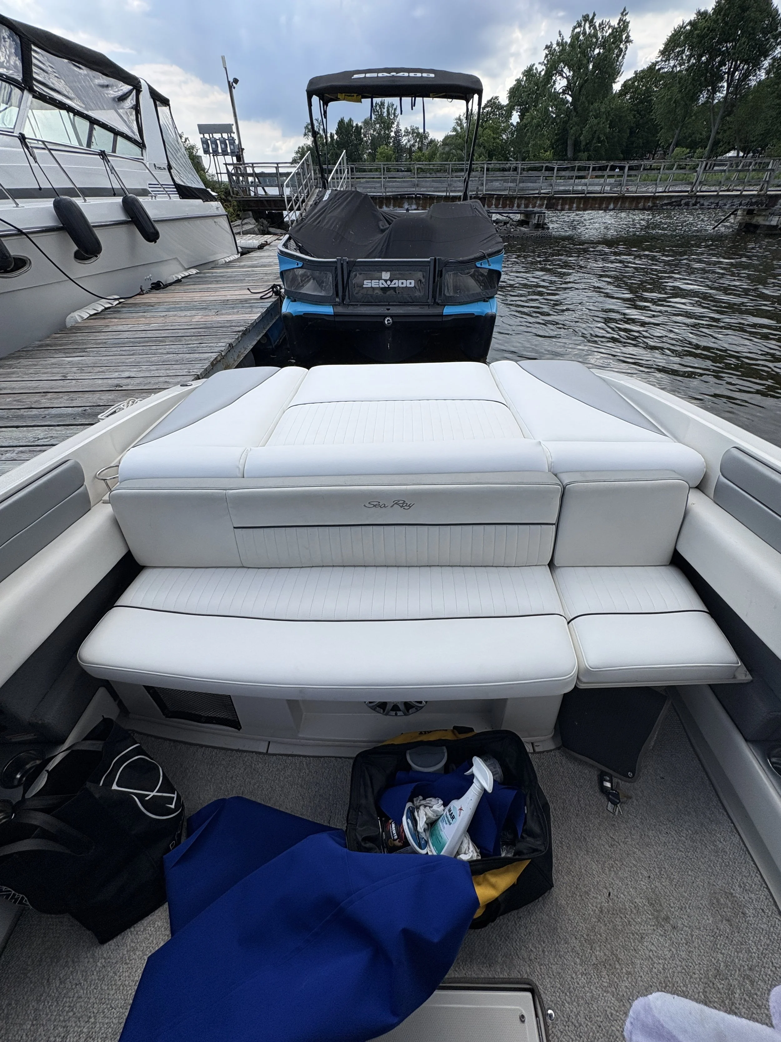 View from the back of a boat showing a white cushioned seating area, a blue duffle bag, and a black bag with toiletries inside. In the background, there are parked boats, a dock, water with gentle ripples, and trees under a partly cloudy sky.