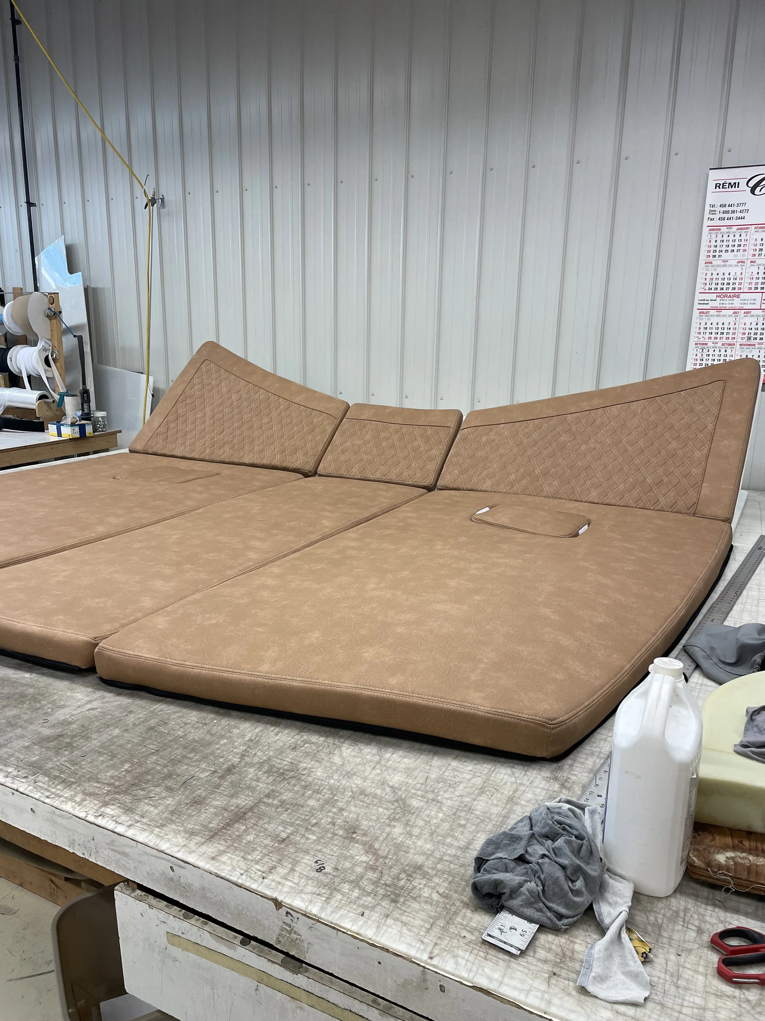 A large brown sectional sofa, with a checkered pattern on the headrest pillows, is situated on a workbench in a workshop or storage area. The workbench has various tools, fabric, and a spray bottle on it. The background features a metal wall with a calendar pinned to it and some electrical cords hanging down.