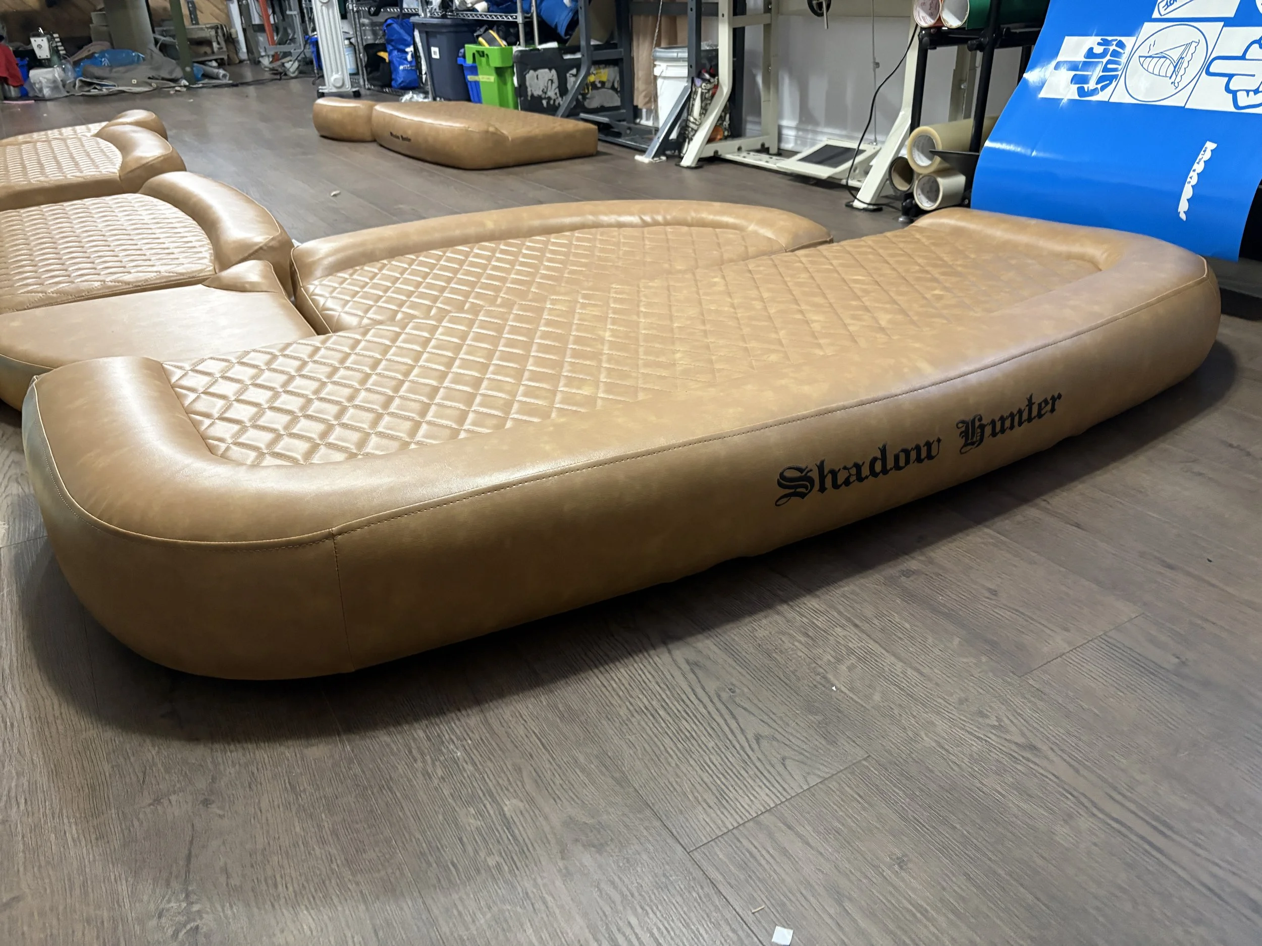 A beige inflatable pool float lying on a wooden floor, with the word 'Shadow Hunter' printed on the side, in a cluttered room with various objects and furniture in the background.