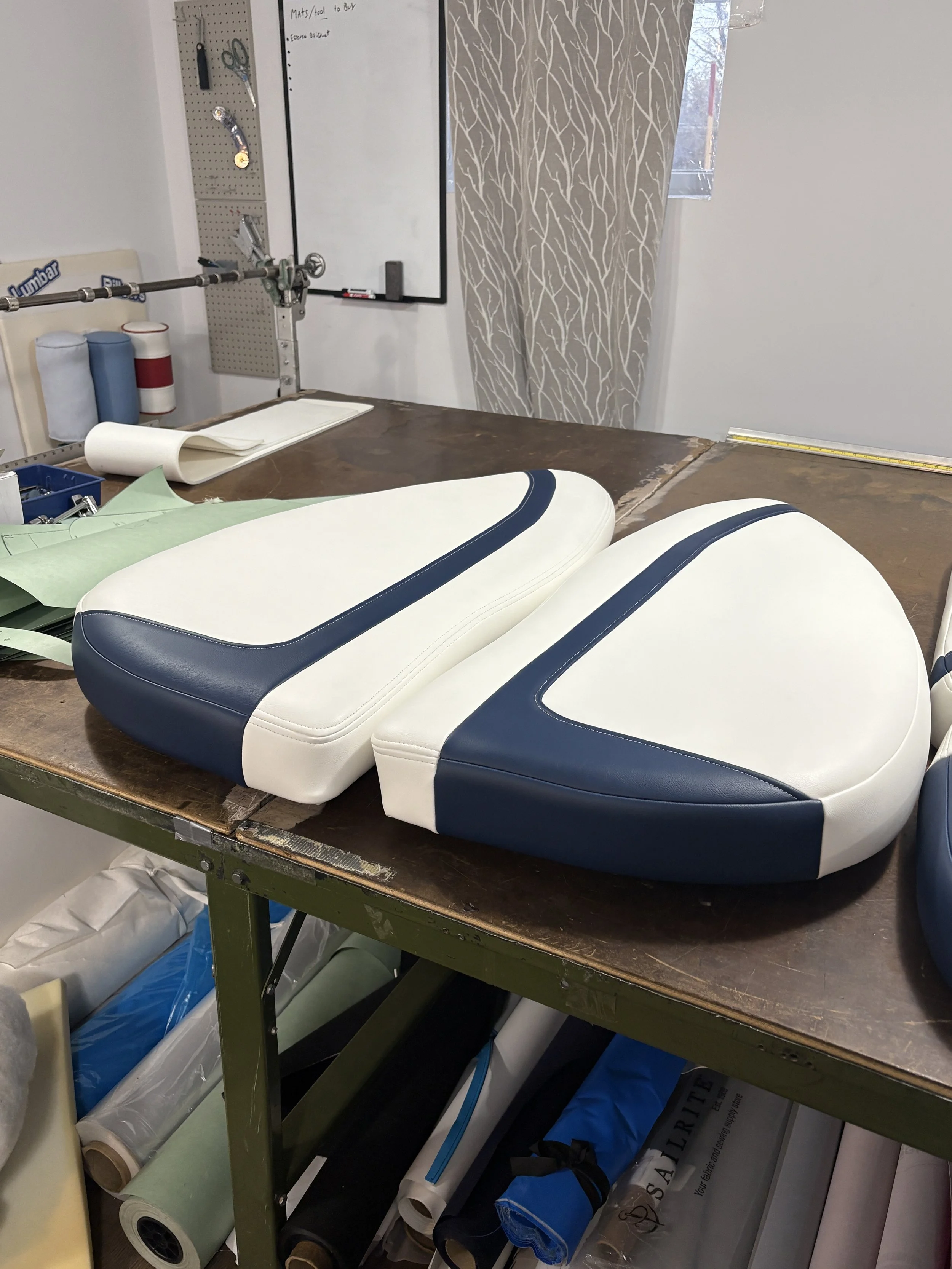 A workbench with a foam or padded material composed of white and navy blue sections, possibly an upholstery or cushioning piece, placed on a wooden table in a workshop or upholstery shop. In the background, there are rolls of fabric or foam, tools hanging on a pegboard, a whiteboard, and a patterned curtain covering part of a window.