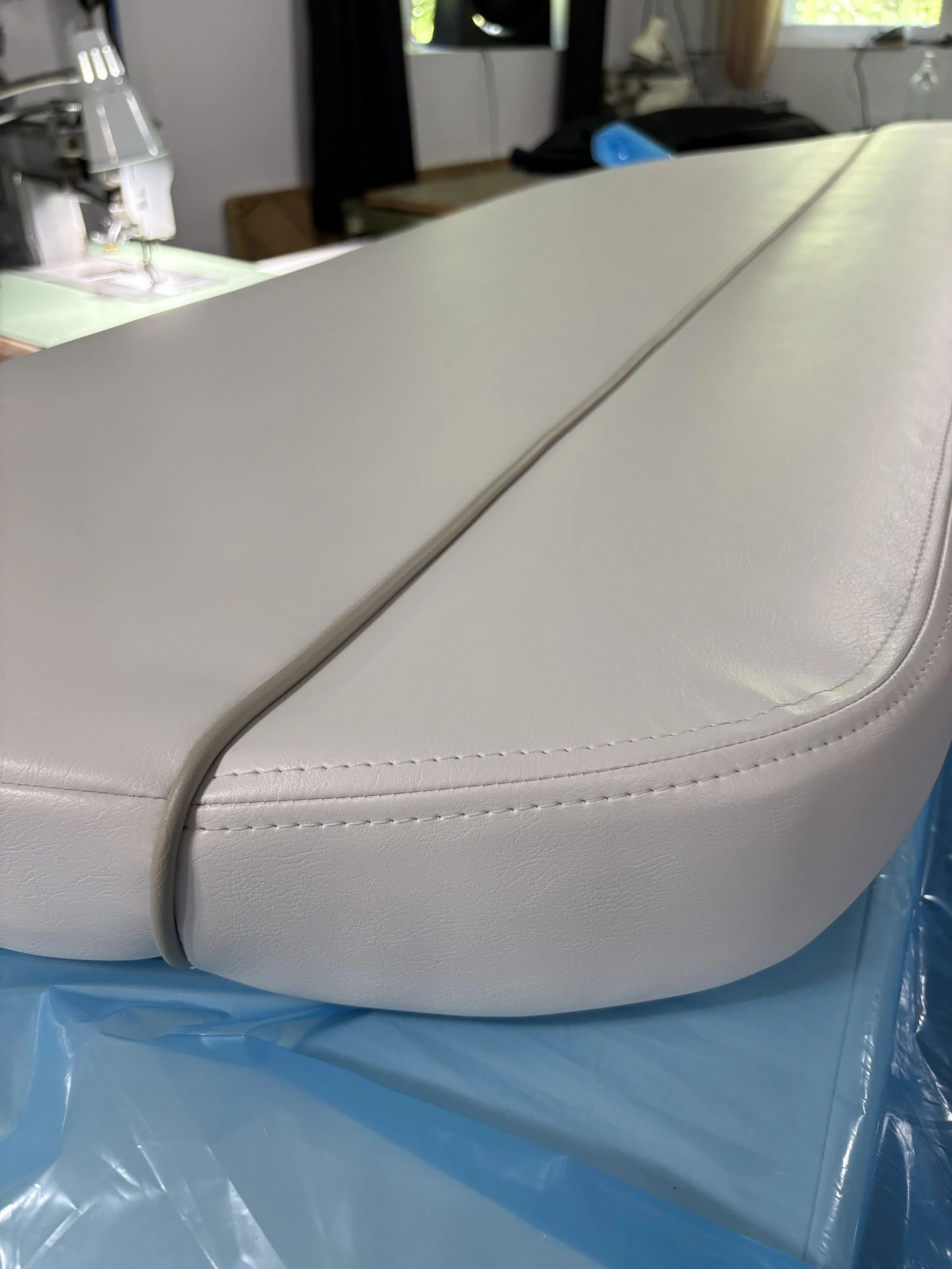 Close-up of a white leather upholstered piece of furniture, possibly a couch or chair, with visible stitching, placed on a blue plastic sheet, and a blurry background of a room with windows and objects.