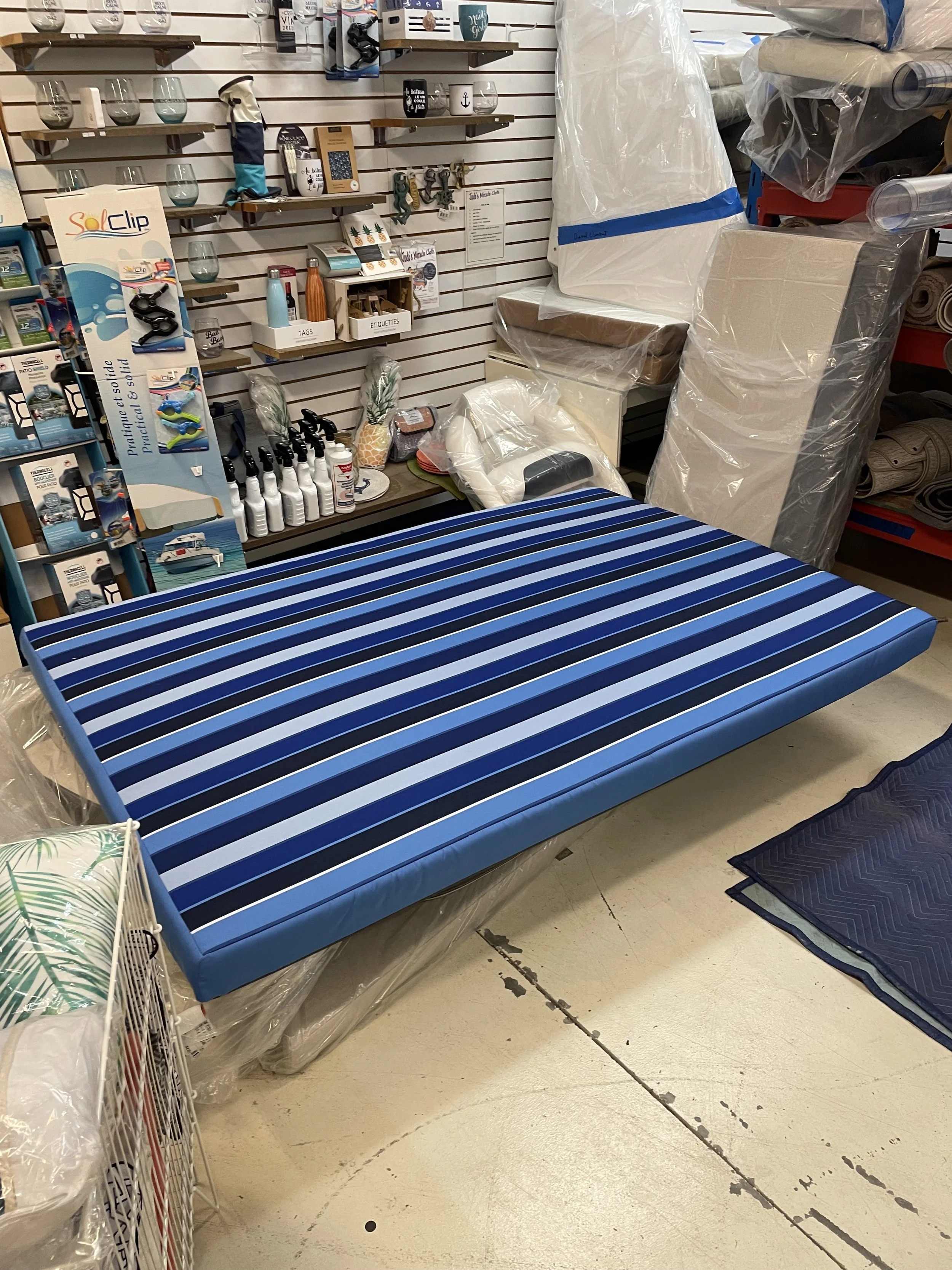 A store display with a blue striped mattress on a metal frame, surrounded by shelves of home goods, including bottles, glasses, towels, and other items.