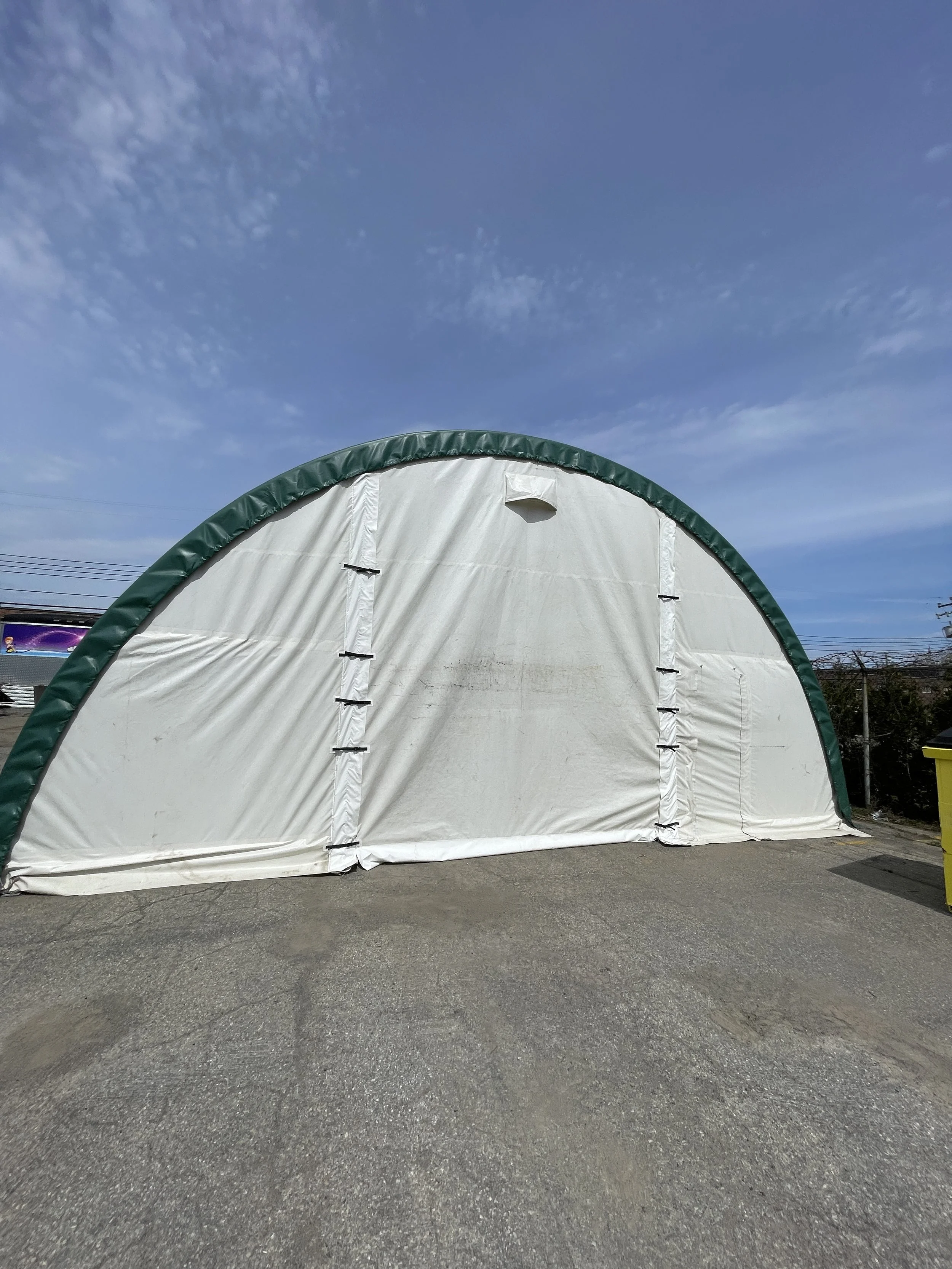 White and green fabric storage tent or shelter with a curved roof, set outdoors on a paved surface under a partly cloudy blue sky.