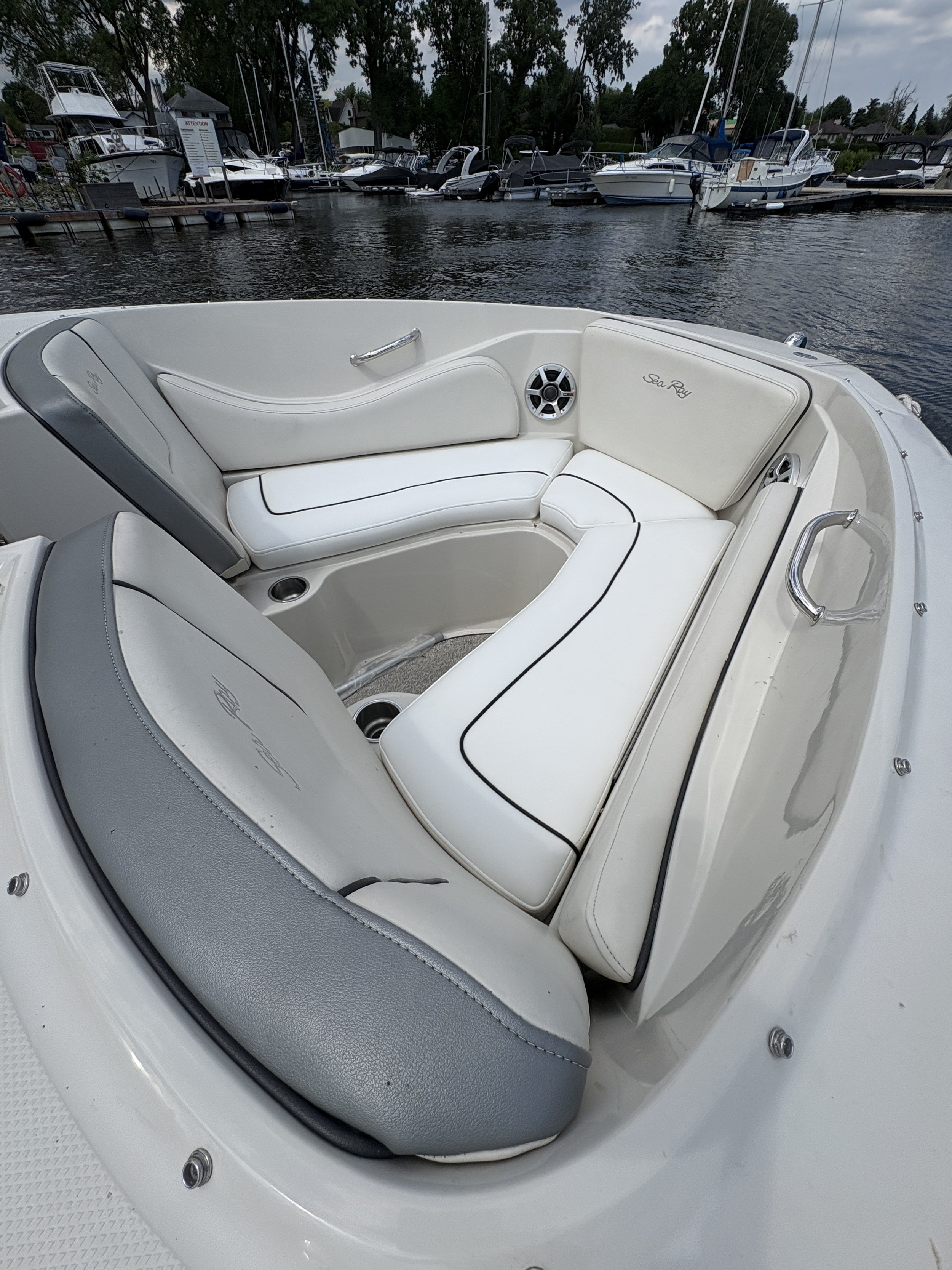 White boat with cushioned seating and a speaker, parked near a marina with other boats and sailboats in the background.
