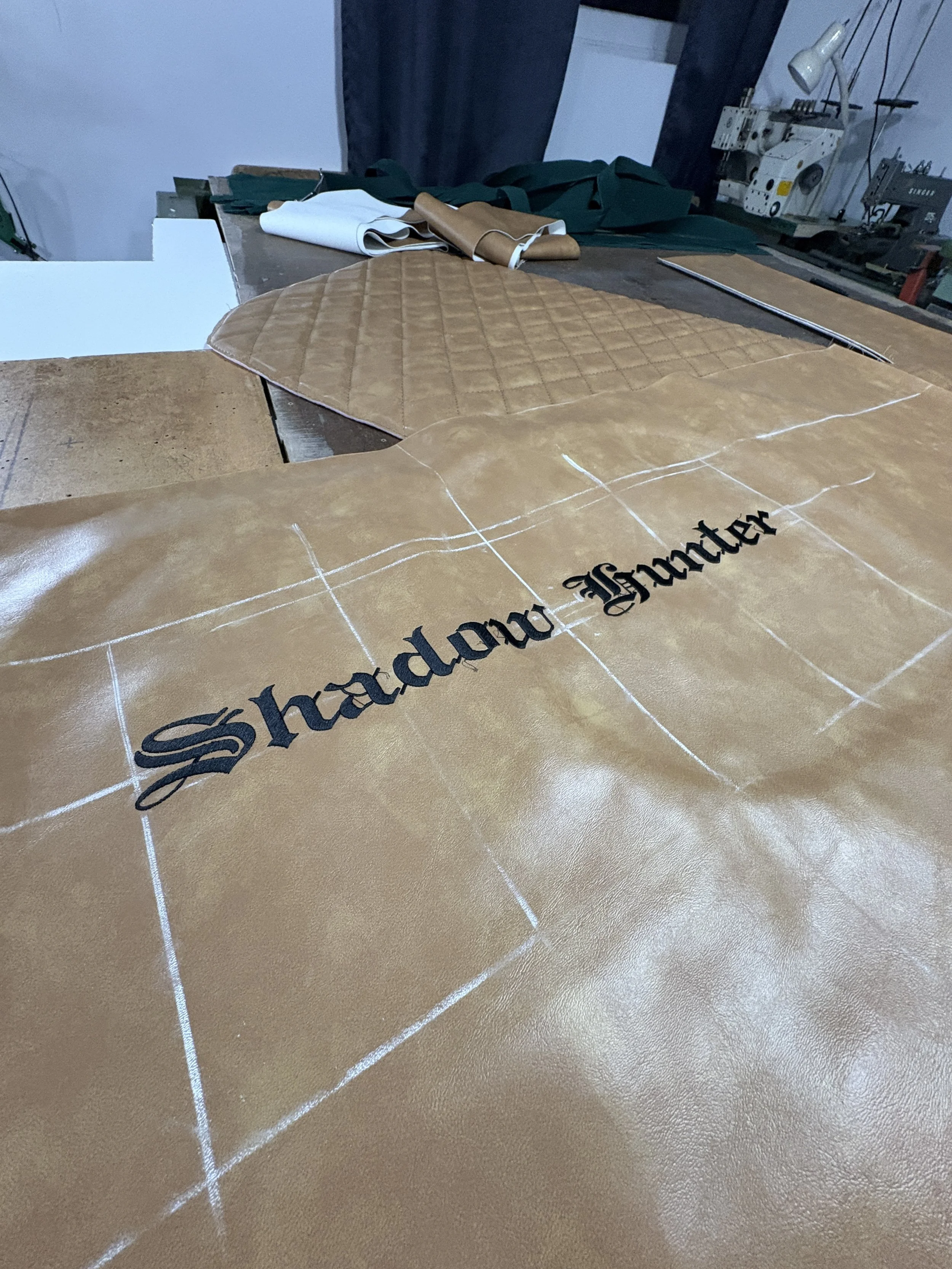 Leather pieces on a worktable with grid lines and the text 'Shadow Box' partially visible, in a workshop setting.
