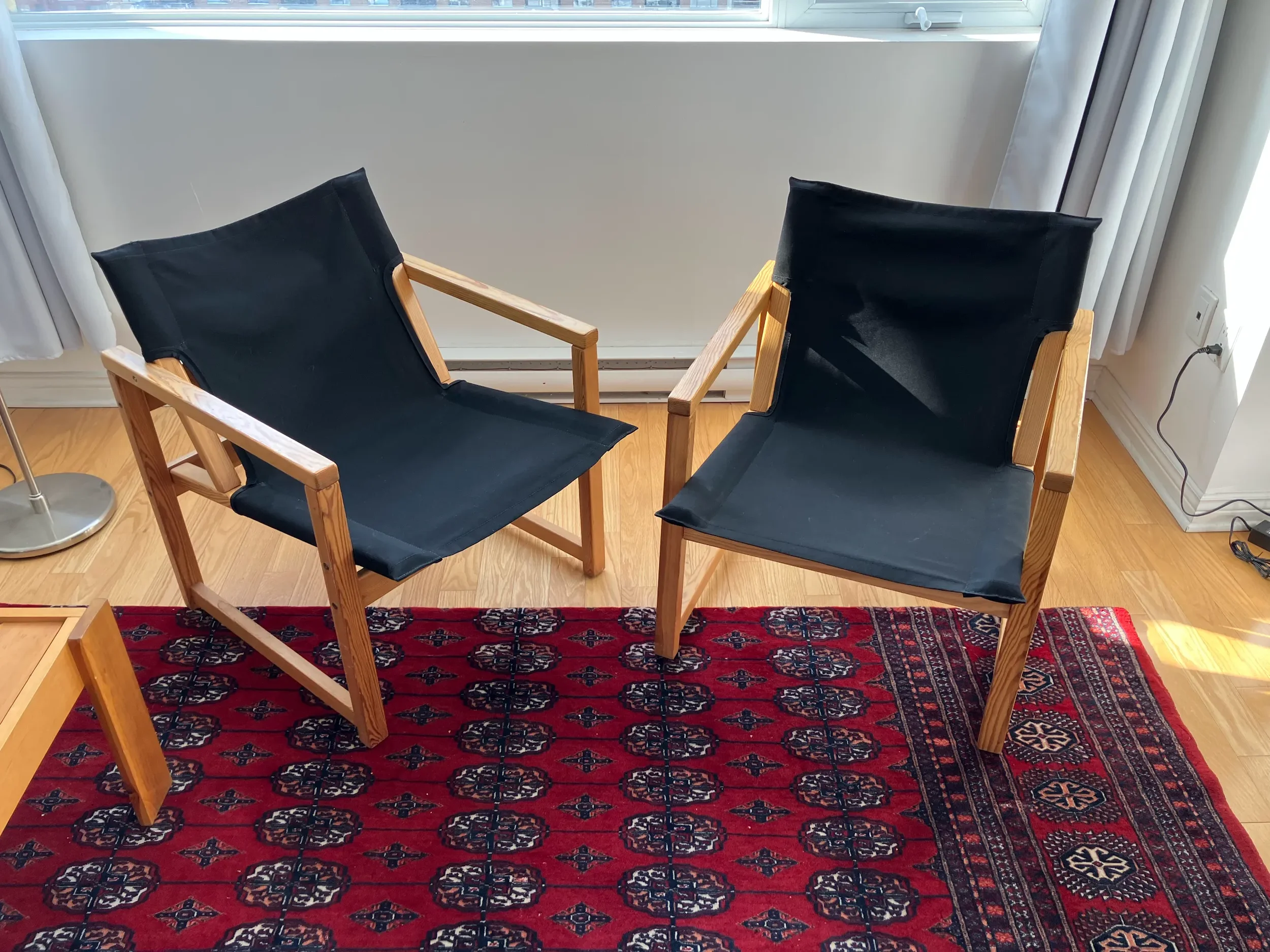 Two wooden chairs with black fabric seats and backs, placed on a red patterned rug near a window with white curtains, in a room with hardwood floors.