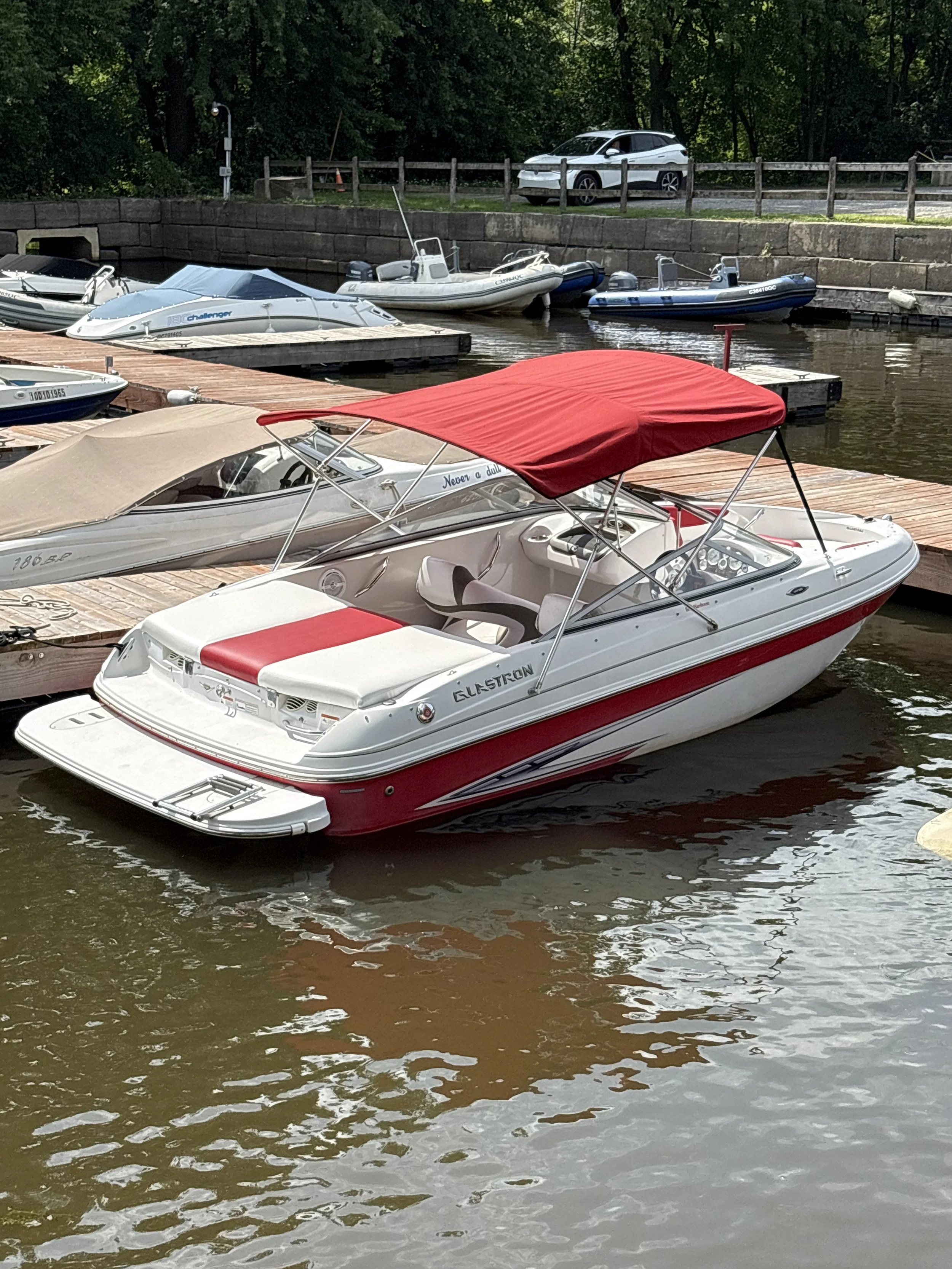 Multiple boats docked at a pier on a river or lake, with a stone retaining wall and parked car in the background, and greenery beyond the fence.