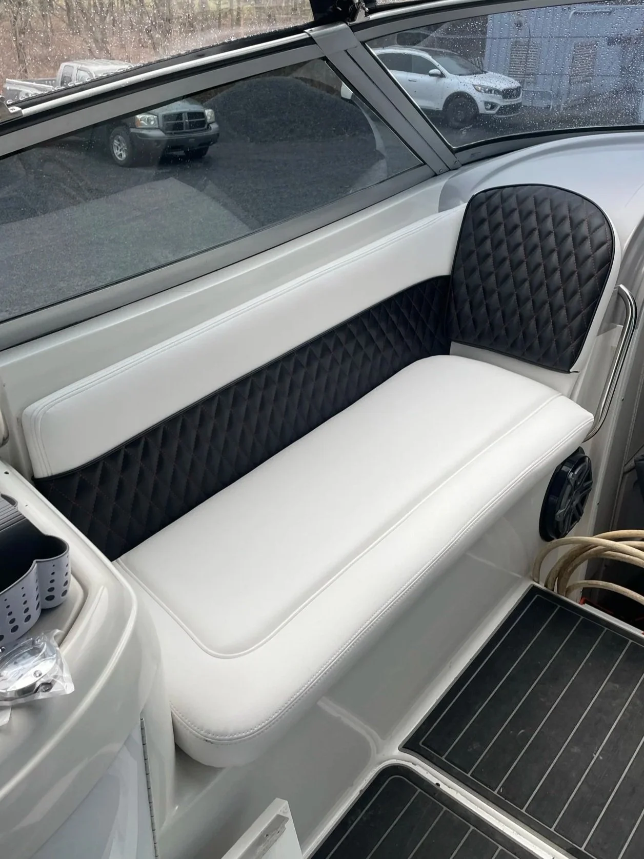 View of the interior of a boat with a built-in seating area. The seating has white cushions with black quilted accents on the backrest. Outside the boat, through a rain-speckled window, there are parked cars visible, including two white vehicles and a gray vehicle.