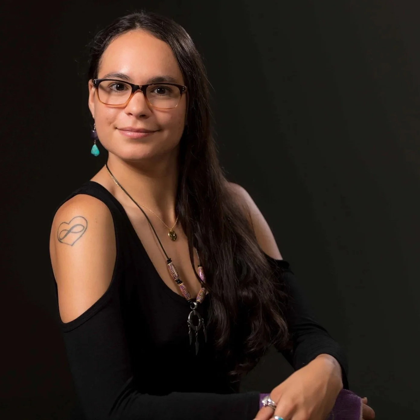 A woman with long dark hair, glasses, and a black top with shoulder cutouts, sitting against a dark background. She has a tattoo on her upper left arm of an infinity symbol within a heart, wears a necklace with beads and a pendant, and has earrings and rings.