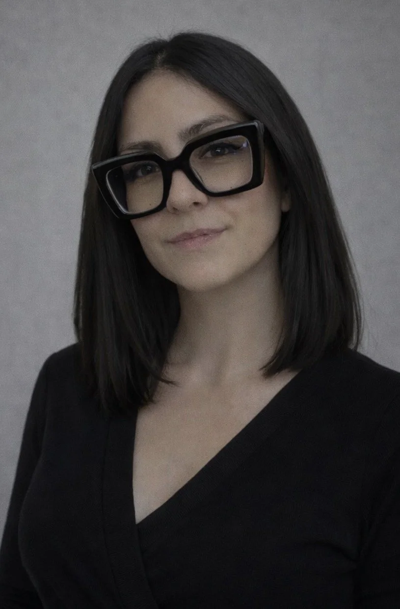 A woman with straight dark hair wearing large black glasses and a black top, standing against a gray background.