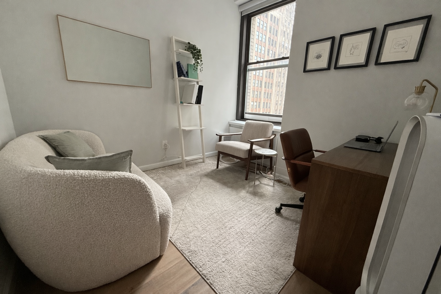 A cozy office with a cream textured sofa with grey pillows, a beige armchair, a wooden desk with a chair, a white ladder bookshelf, a window showing buildings outside, and a wall with the minimalist framed art.