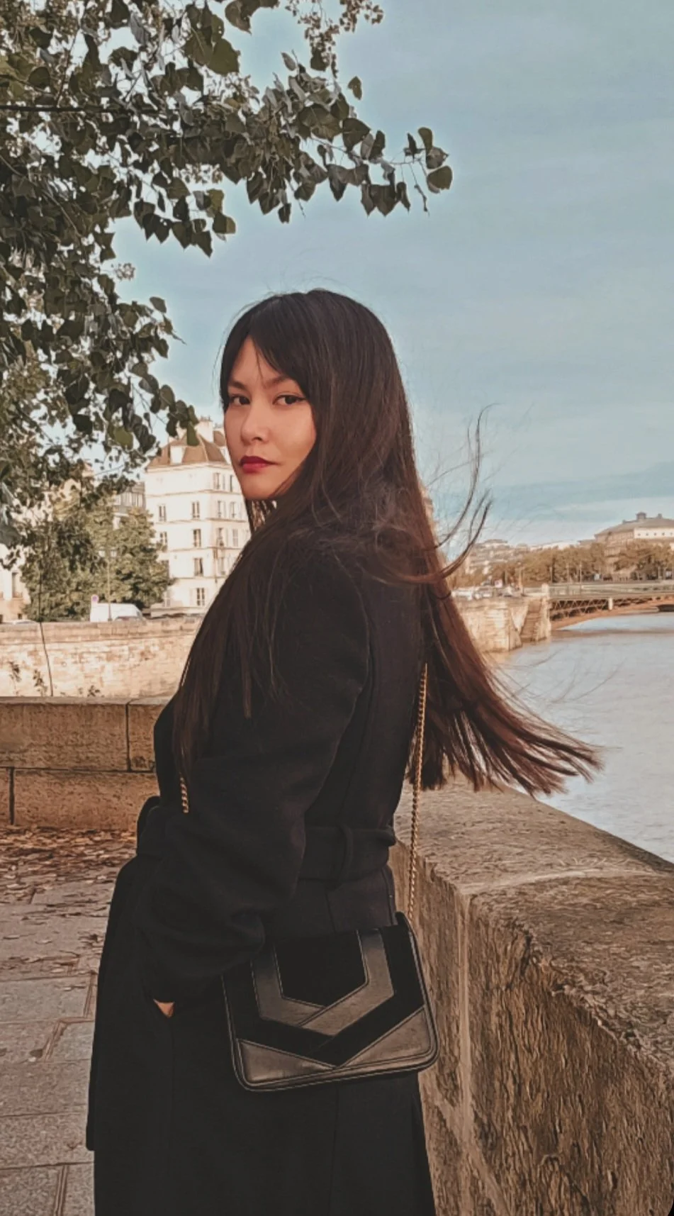A woman with long dark hair stands by a stone railing by the Seine in Paris, with Haussmannian buildings and trees in the background, looking at the camera.