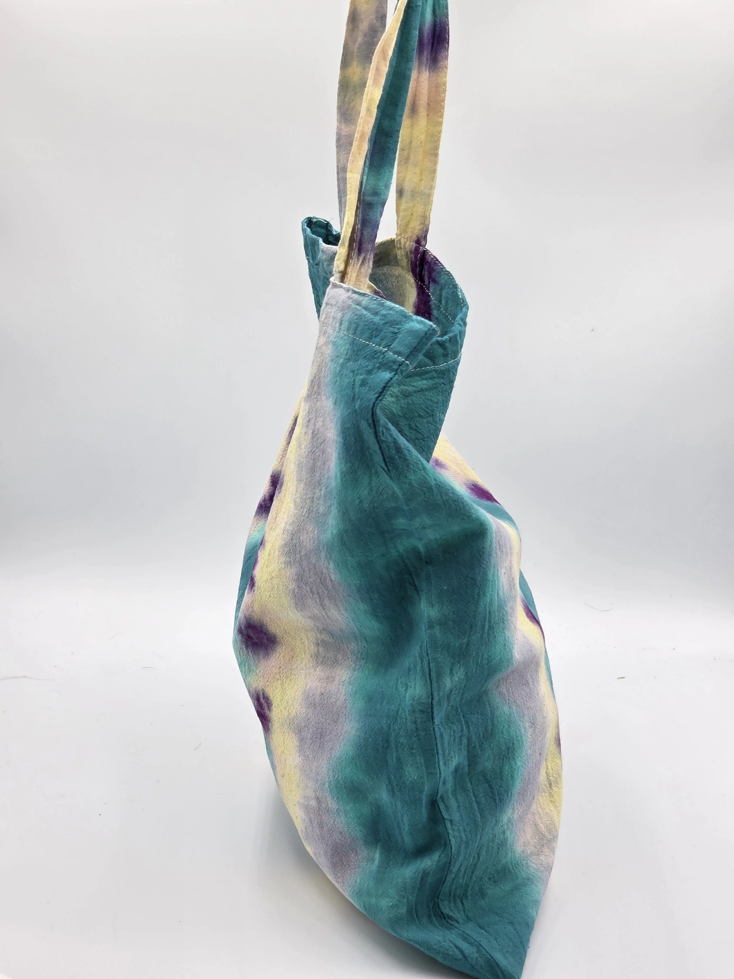 A calico tote bag featuring vertical tie-dye stripes of teal, yellow, and lavender with purple splotches along the center.