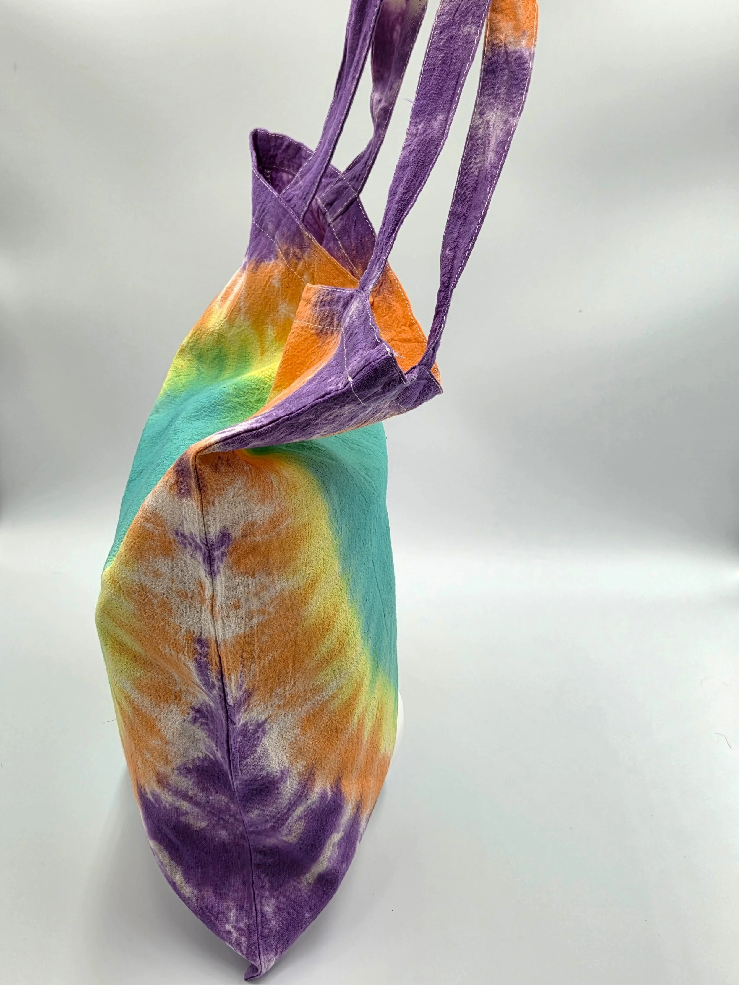 A calico tote bag featuring a tie-dye pattern with a large teal oval center surrounded by glowing rings of yellow, orange, and purple.