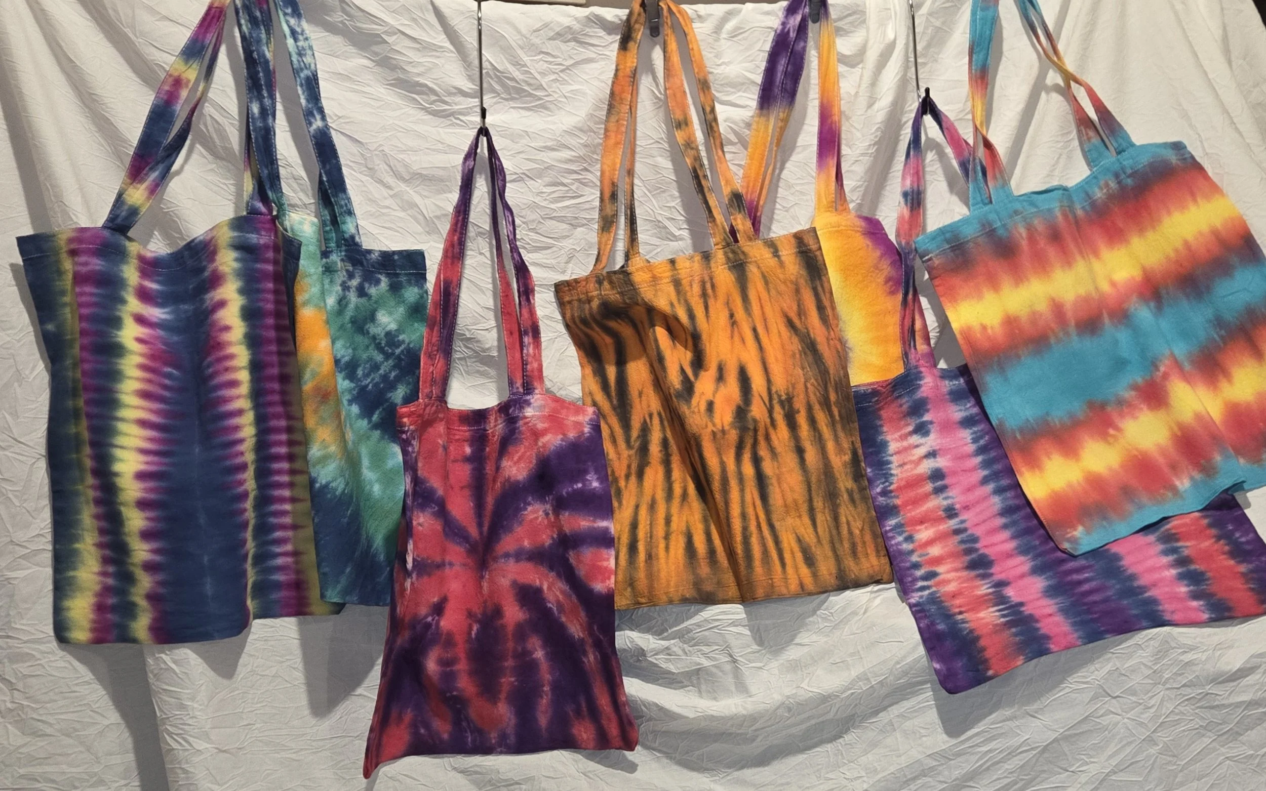 Six colorful tie-dye tote bags hanging against a white background in various vibrant patterns and colors.