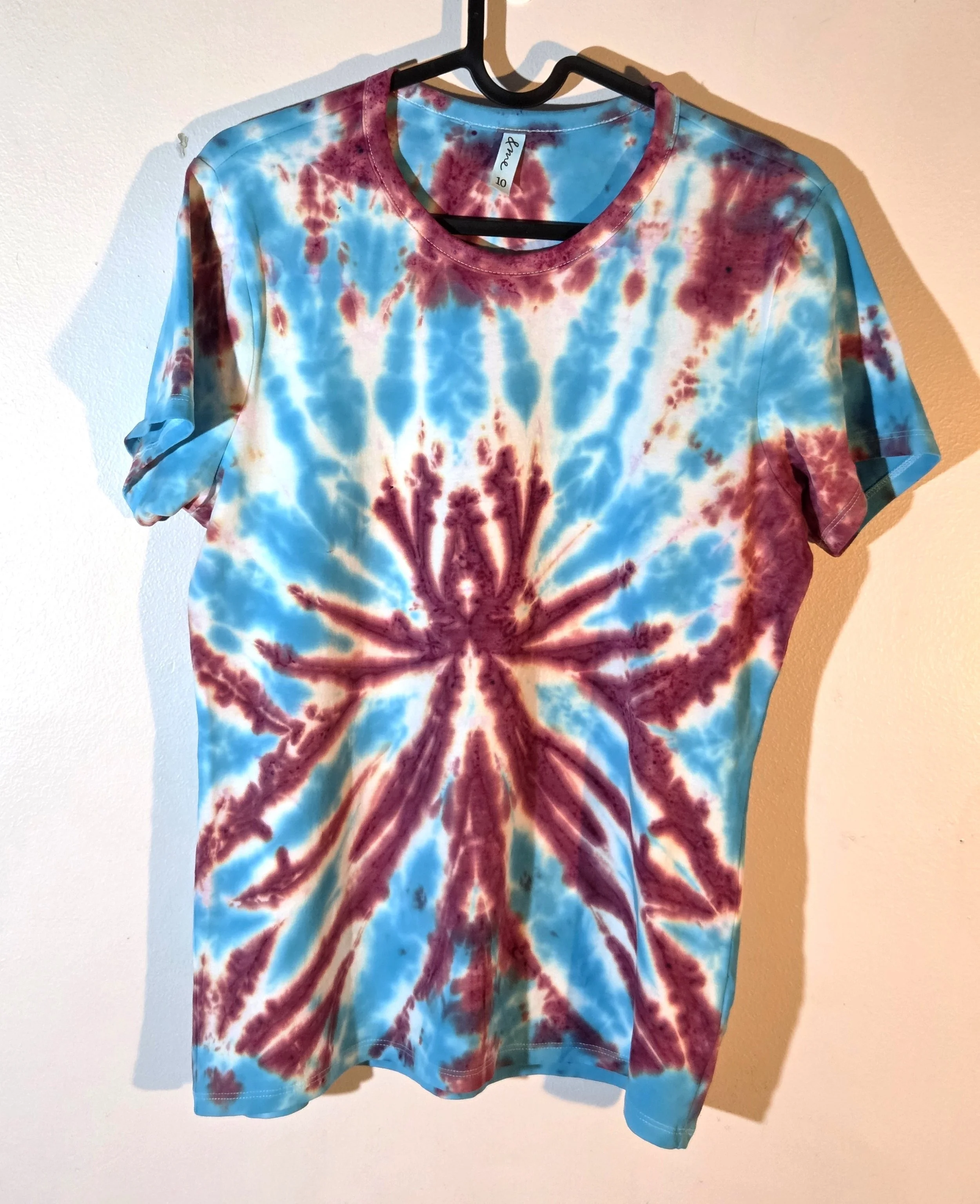 A size 10 t-shirt featuring a tie-dye pattern of a sharp, spiky purple and wine flower on a sky blue and white background.